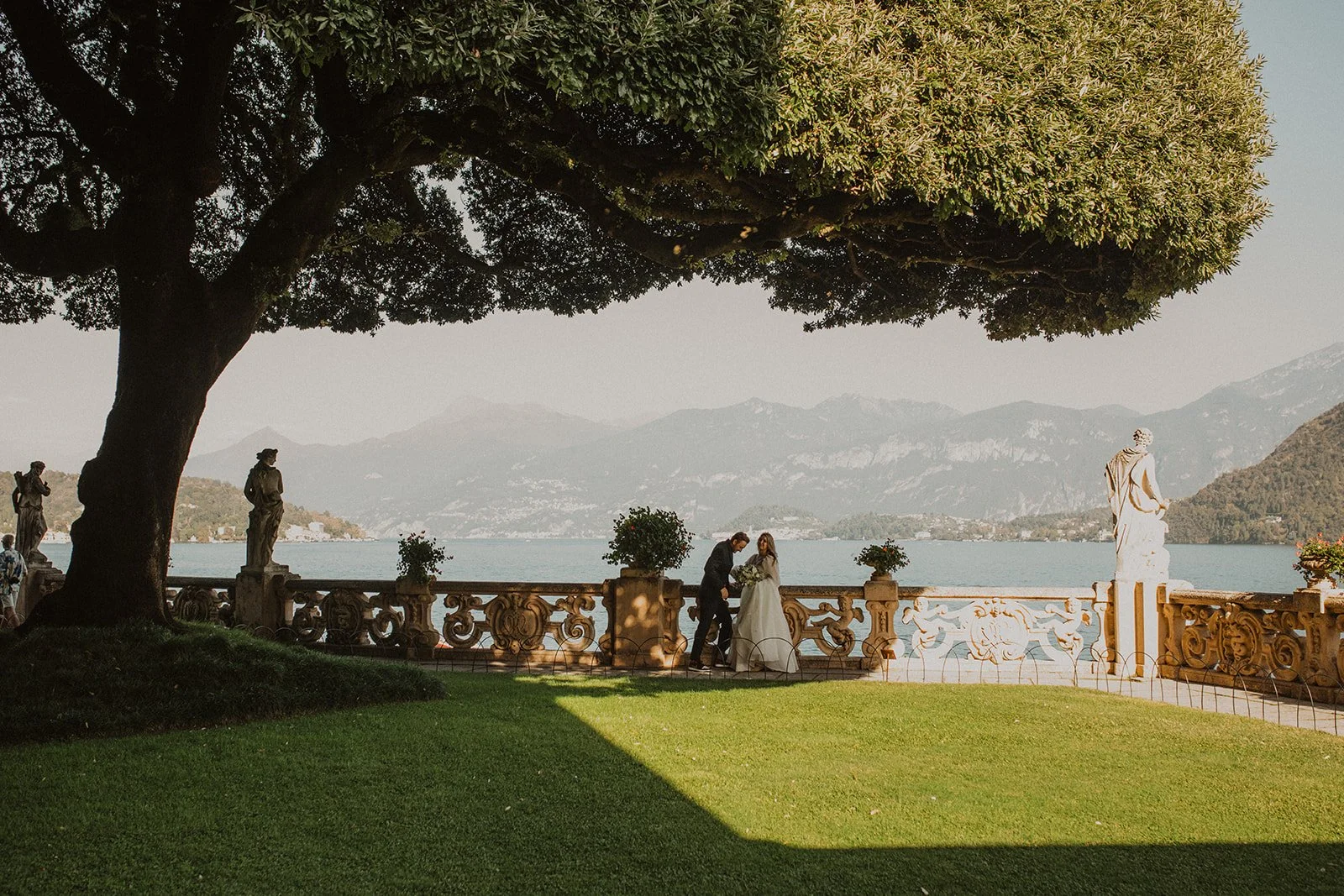 Bride and groom walking through the gardens at Villa del Balbianello during relaxed wedding portraits with Lake Como and the surrounding mountains in the background.