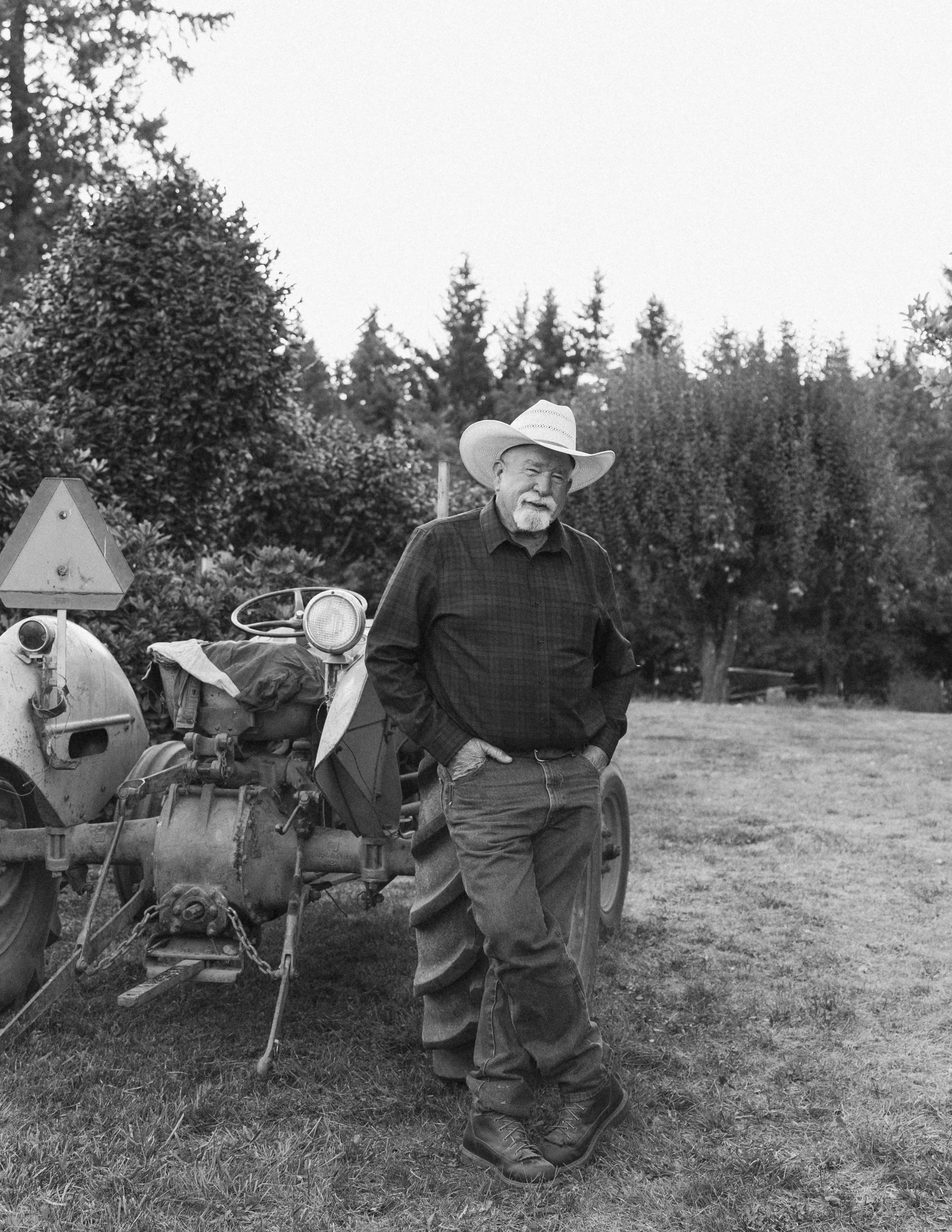 Family Session in Aurora, Oregon on the Koch Family Farm. An elderly man standing outdoors next to an old tractor, wearing a cowboy hat, casual shirt, jeans, and boots, with trees in the background.