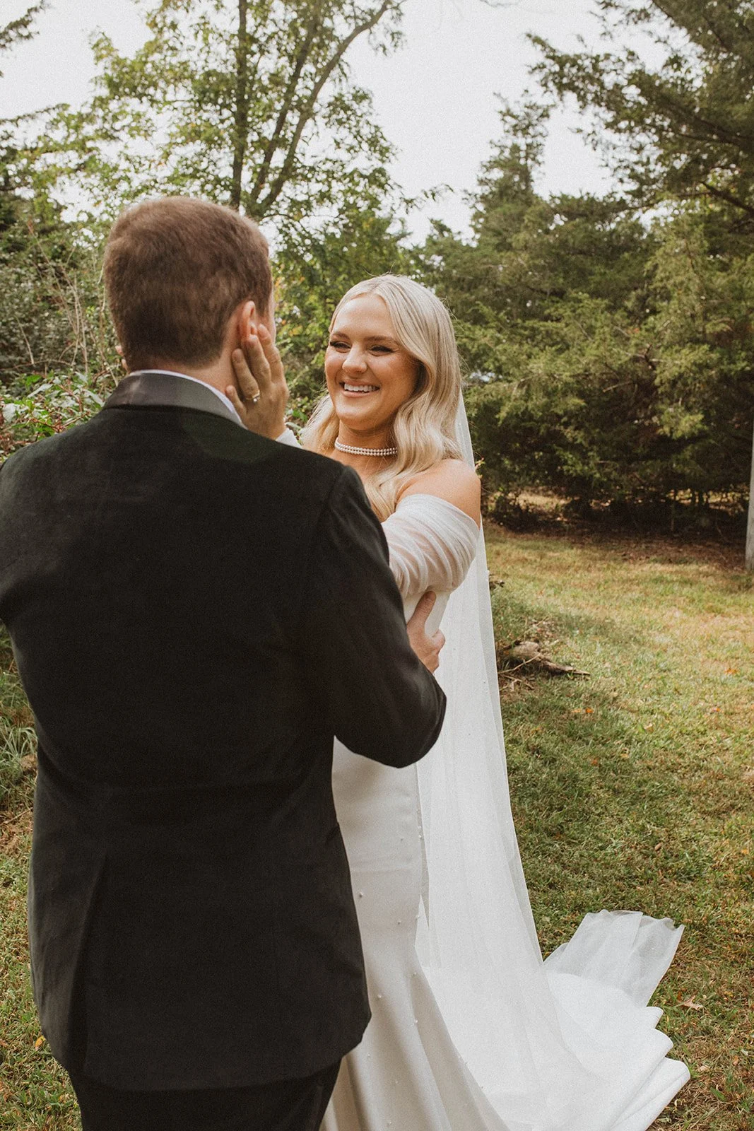 A bride with blonde hair in a white wedding gown smiling at a groom with brown hair in a black tuxedo, outdoors surrounded by trees during a wedding ceremony.