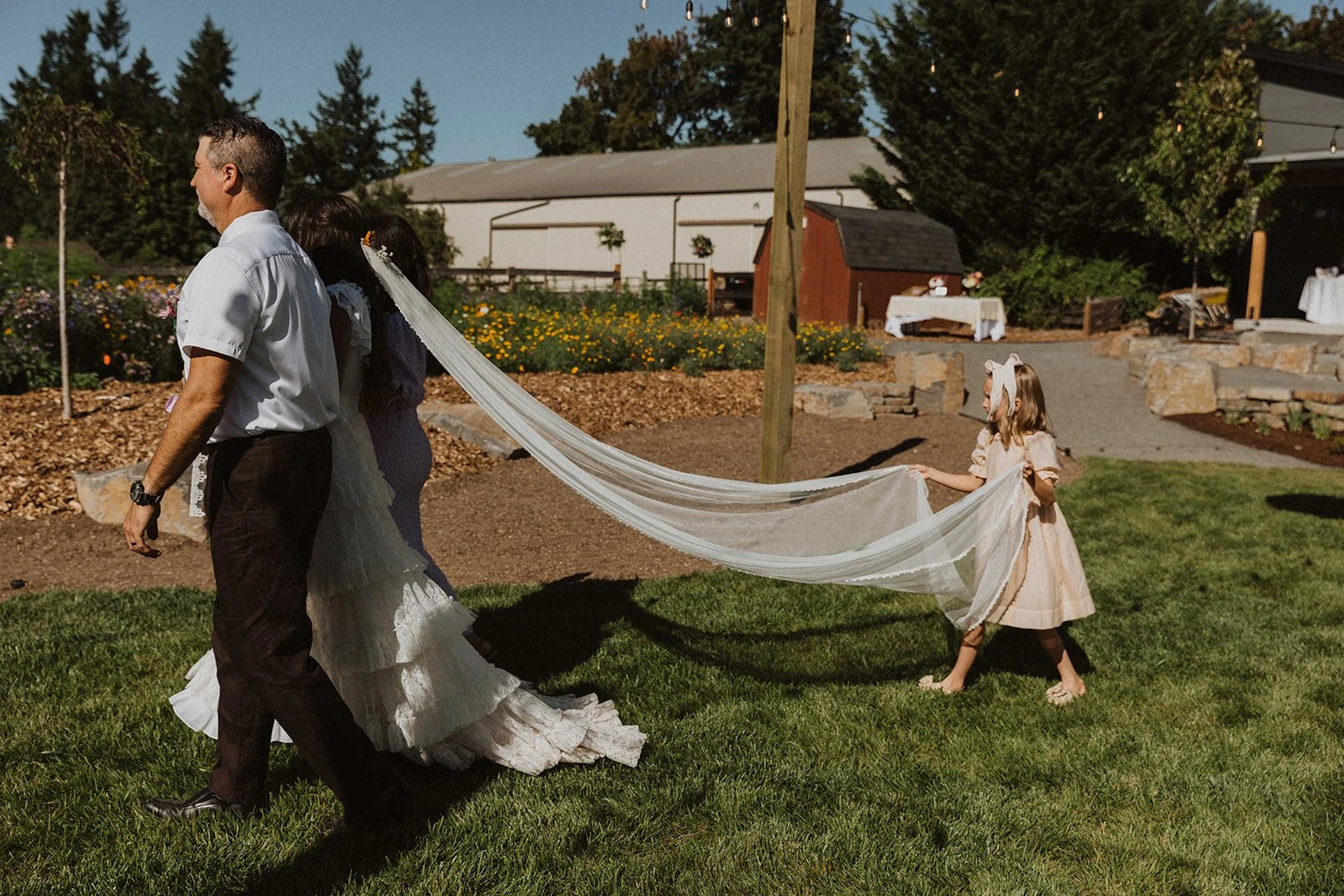 Bride walking toward the ceremony with her veil trailing behind, assisted by a young flower girl holding the veil in an outdoor garden setting