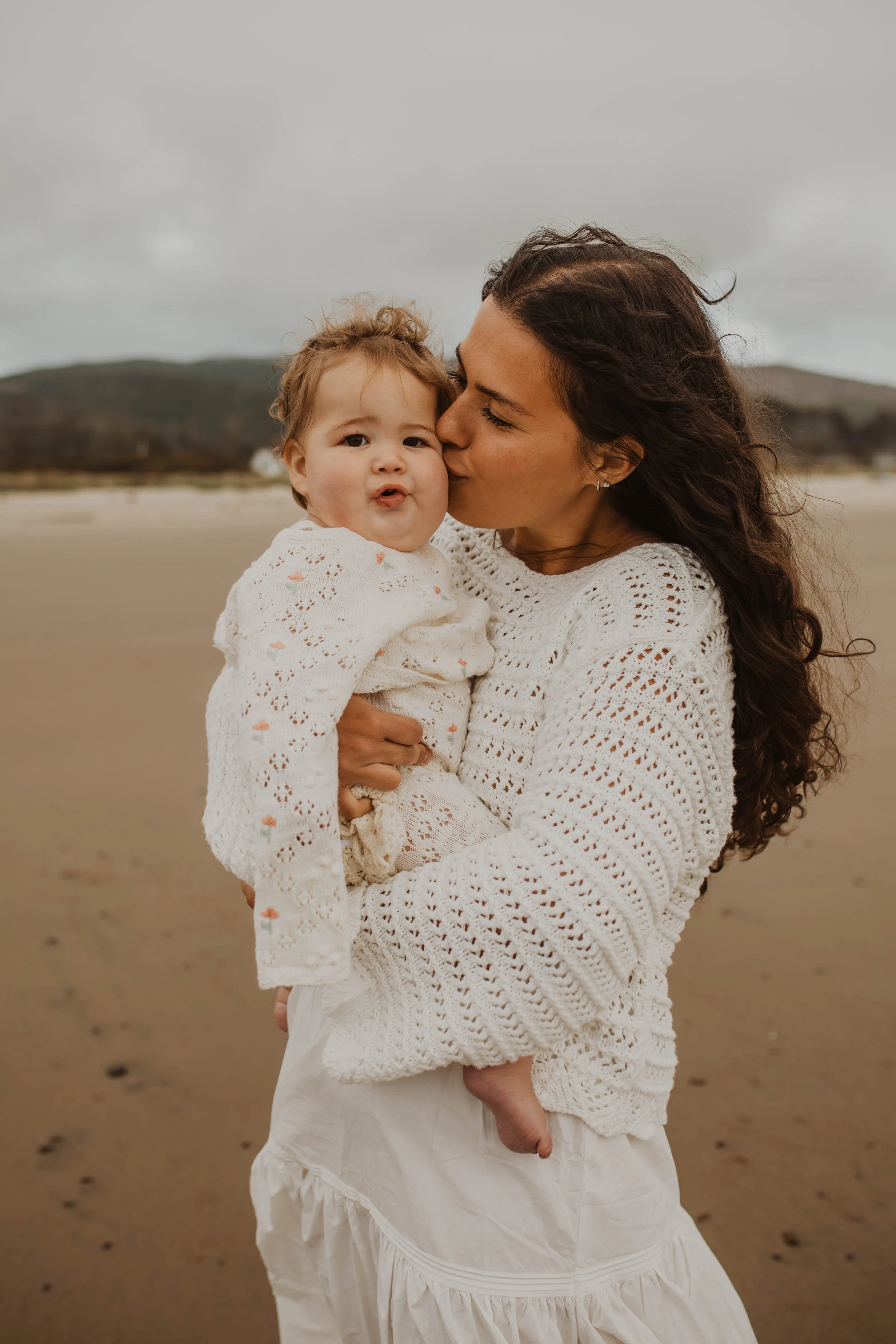 A woman with long brown curly hair kissing a toddler girl on the cheek at the beach on a cloudy day.
