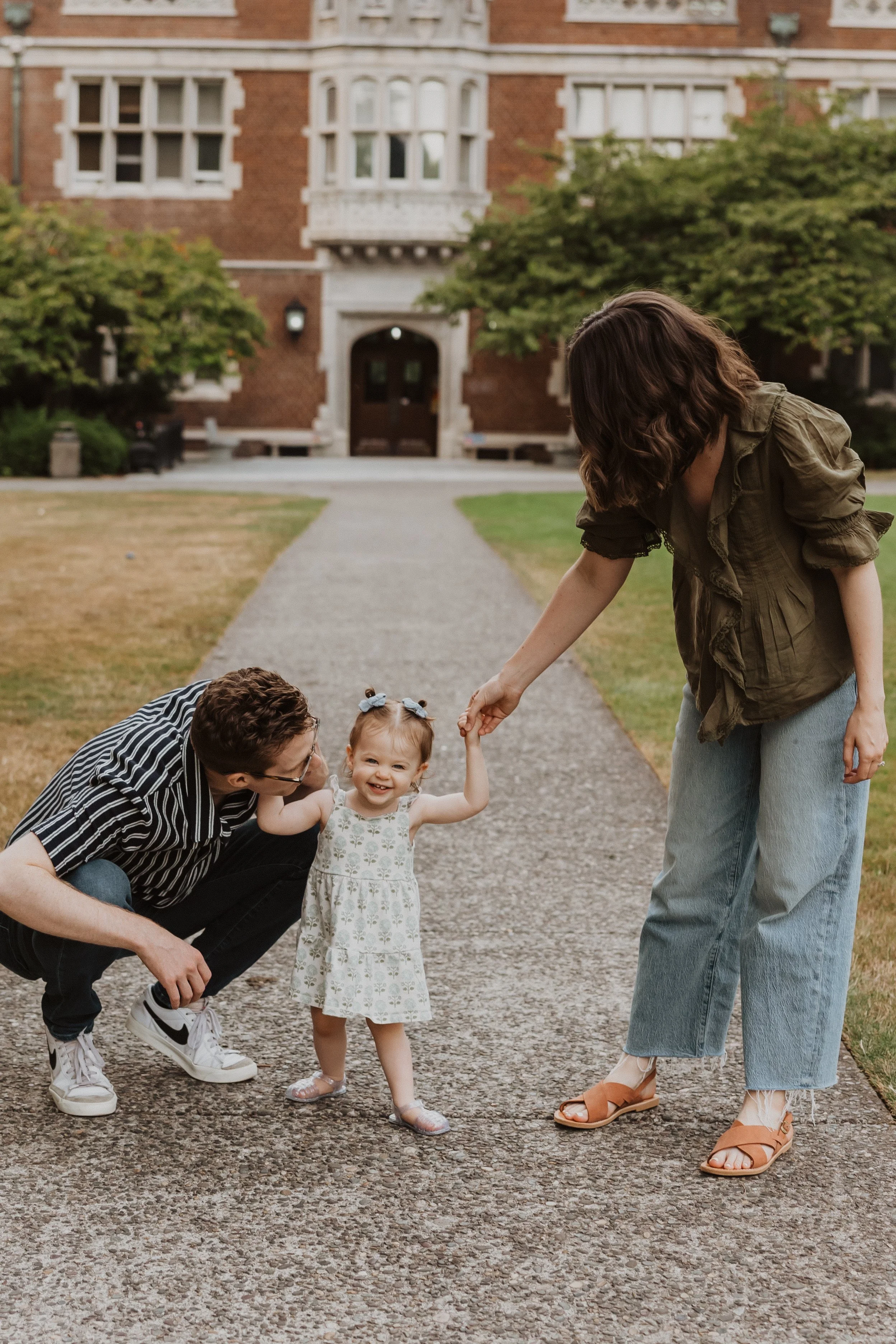 A young girl in a white dress with blue bows in her hair is smiling and holding hands with two women, one squatting on the left and another standing on the right, on a sidewalk in front of a red brick building with greenery.