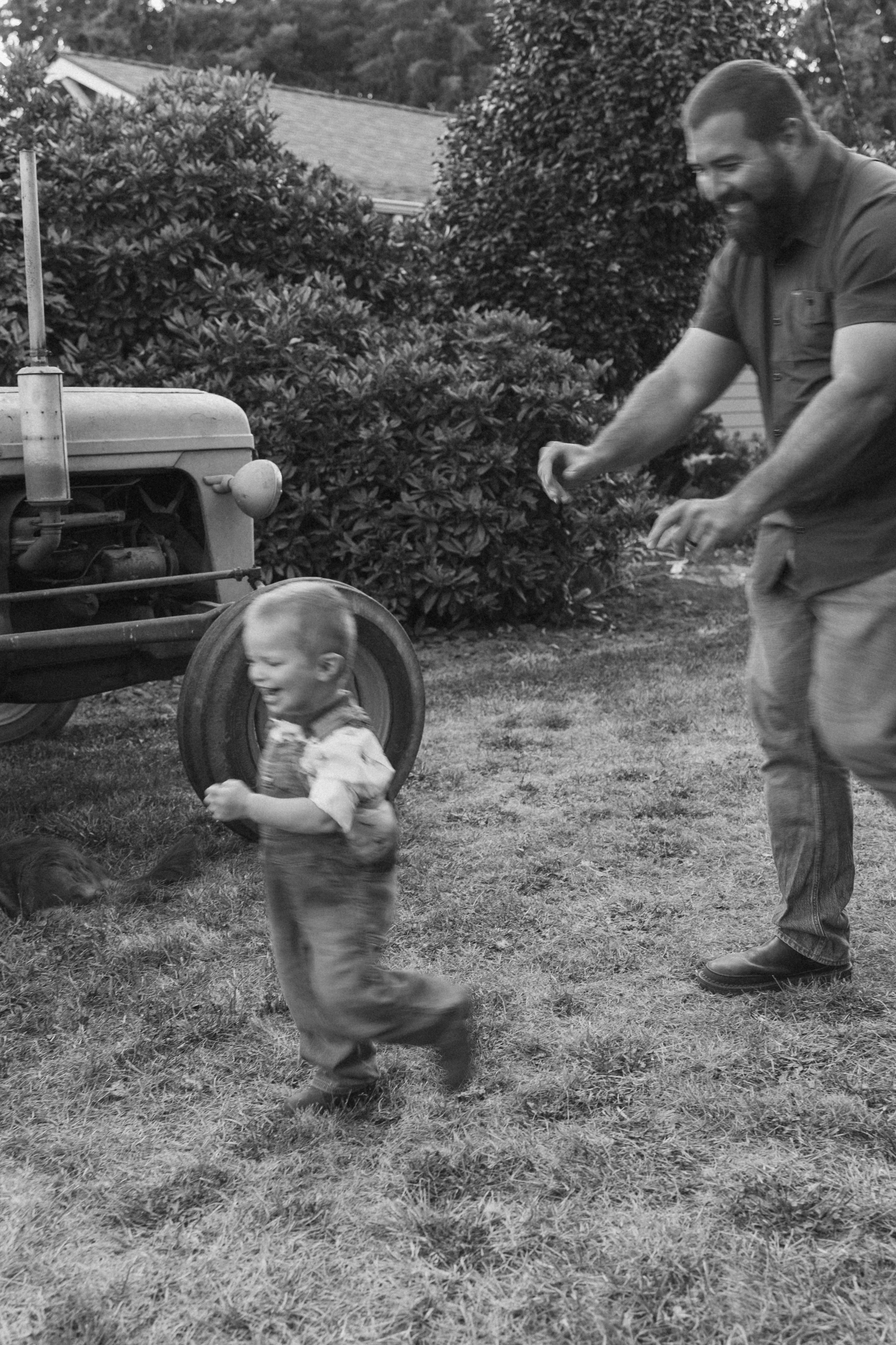 Family Session in Aurora, Oregon on the Koch Family Farm. A smiling young boy running on a grassy yard with an older man, possibly his father, in the background. There is an old tractor and bushes in the backyard, all in black and white.