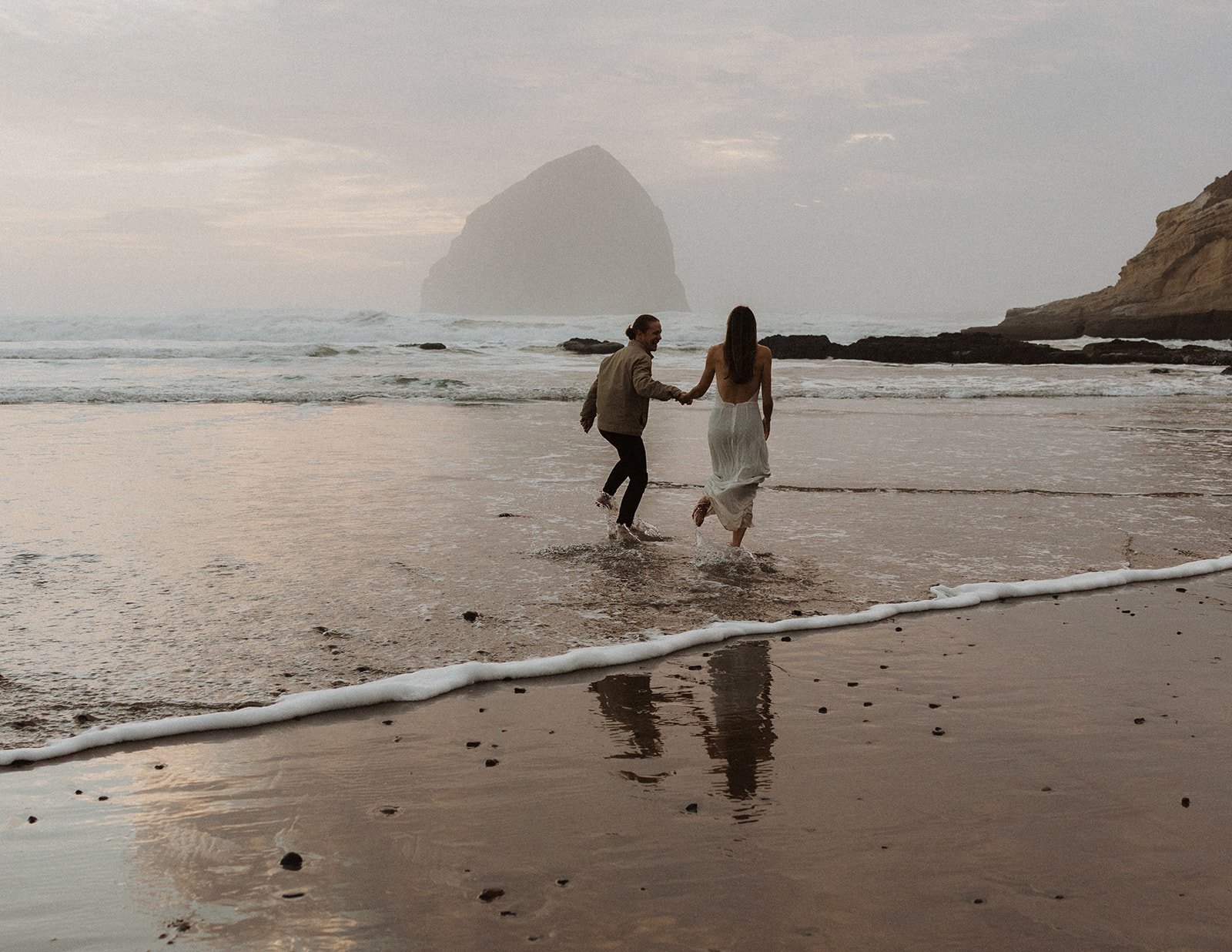 Couple run into the surf together during their Oregon beach engagement session at Cape Kiwanda.