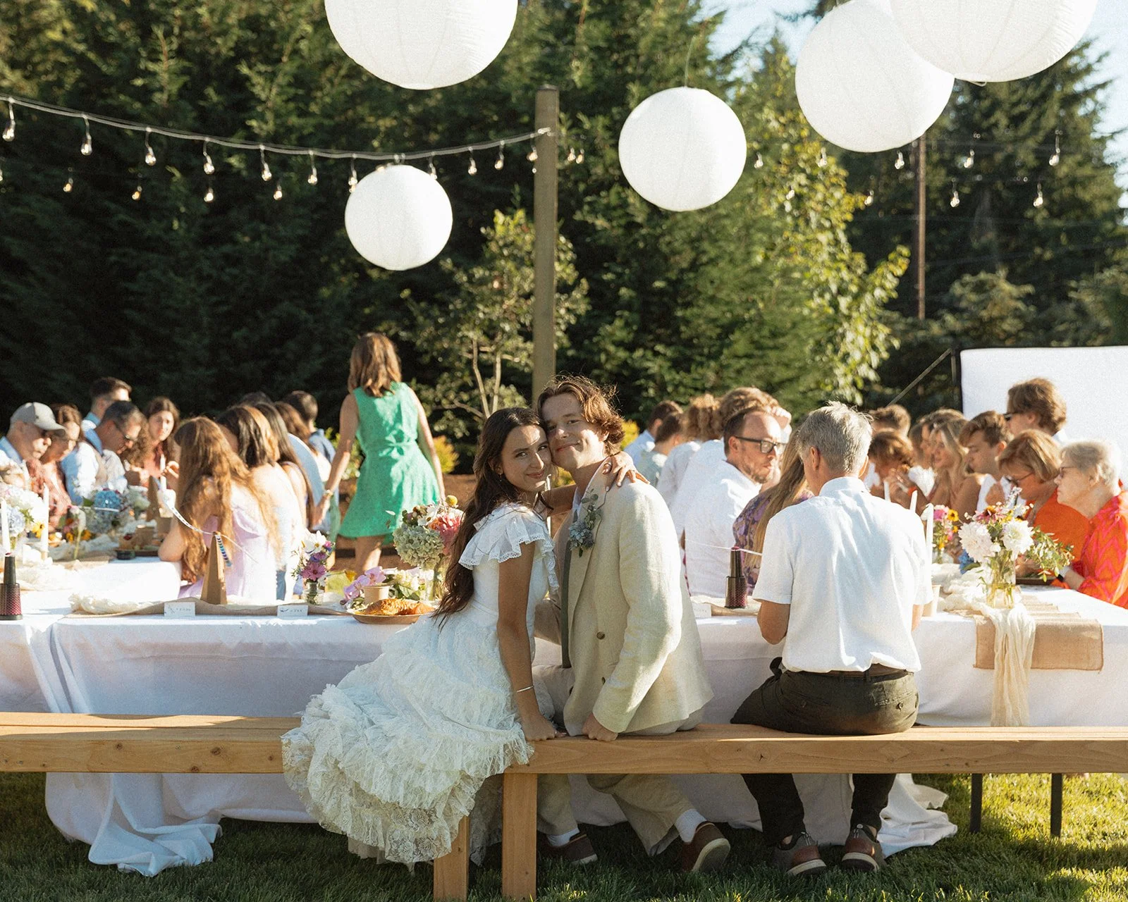 Newlywed couple seated at long outdoor reception table with guests under string lights and lanterns