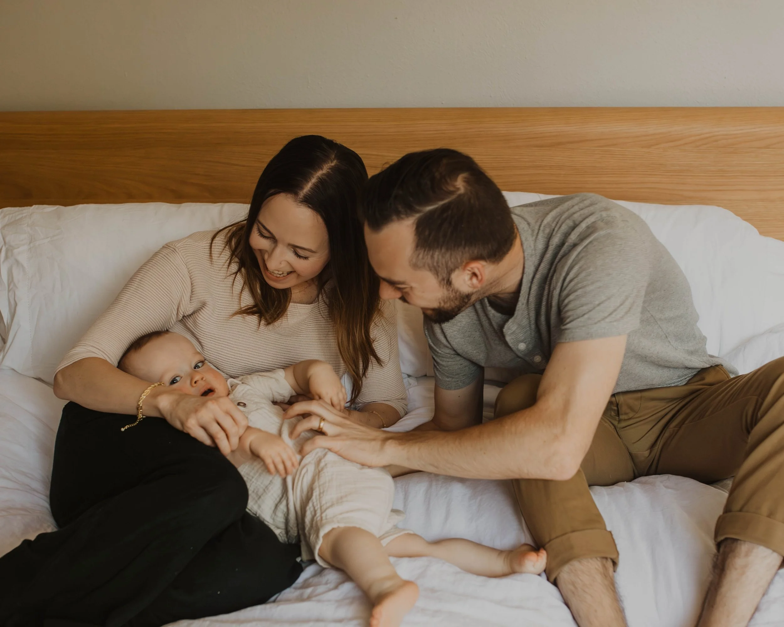 A happy family, including a woman, a man, and a baby, sitting on a bed laughing and playing together.