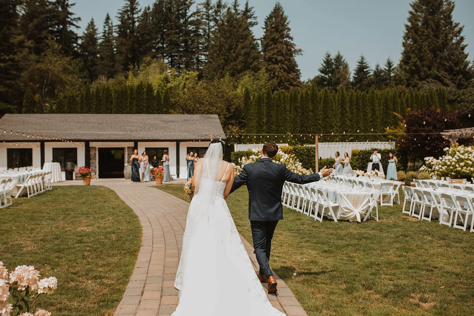 Newlywed couple walking hand in hand into their outdoor wedding reception space with long reception tables, string lights, and guests waiting in the background.