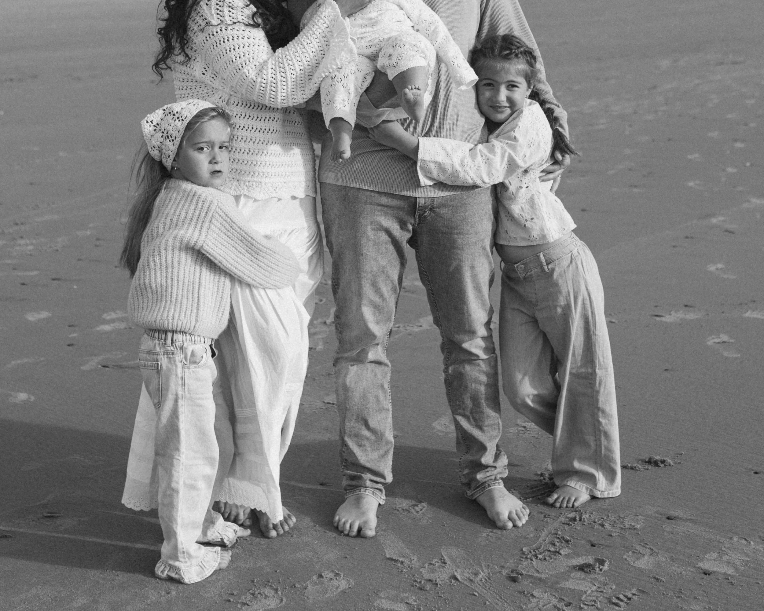 Group of children and an adult standing barefoot on the beach, with a girl hugging the adult's leg and another girl standing nearby, all dressed in casual clothing.