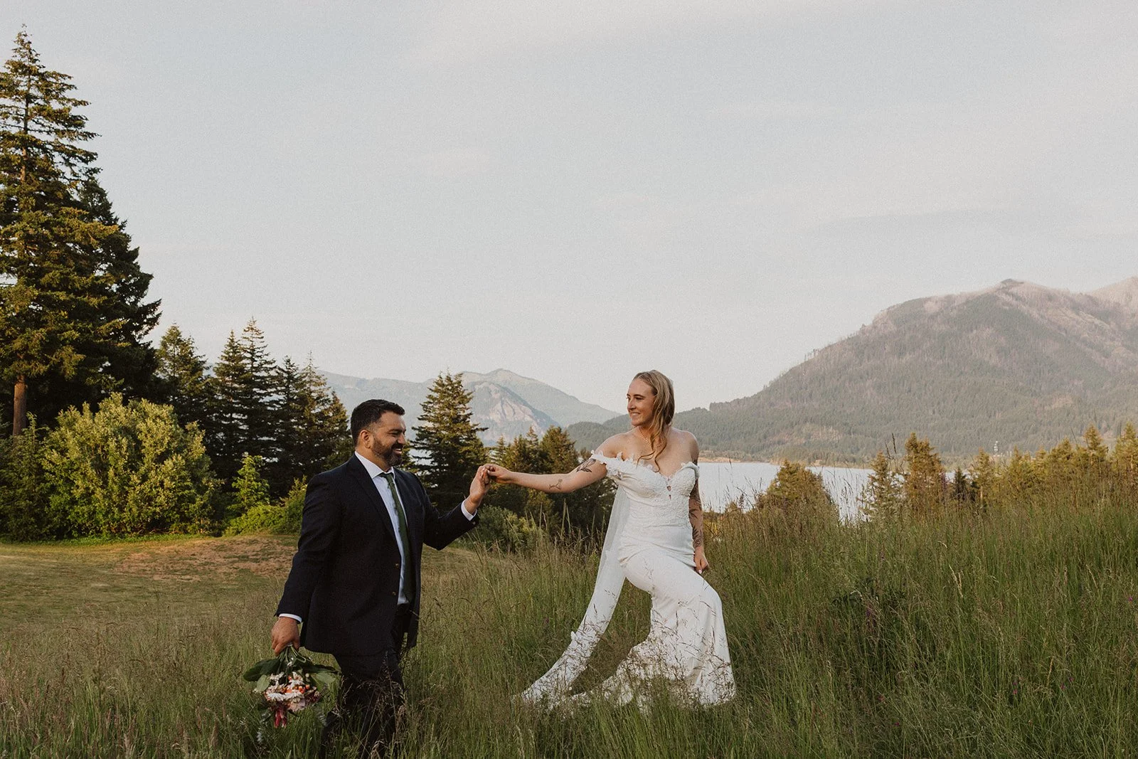 Couple walking hand in hand through a meadow with Columbia River Gorge scenery in the background.
