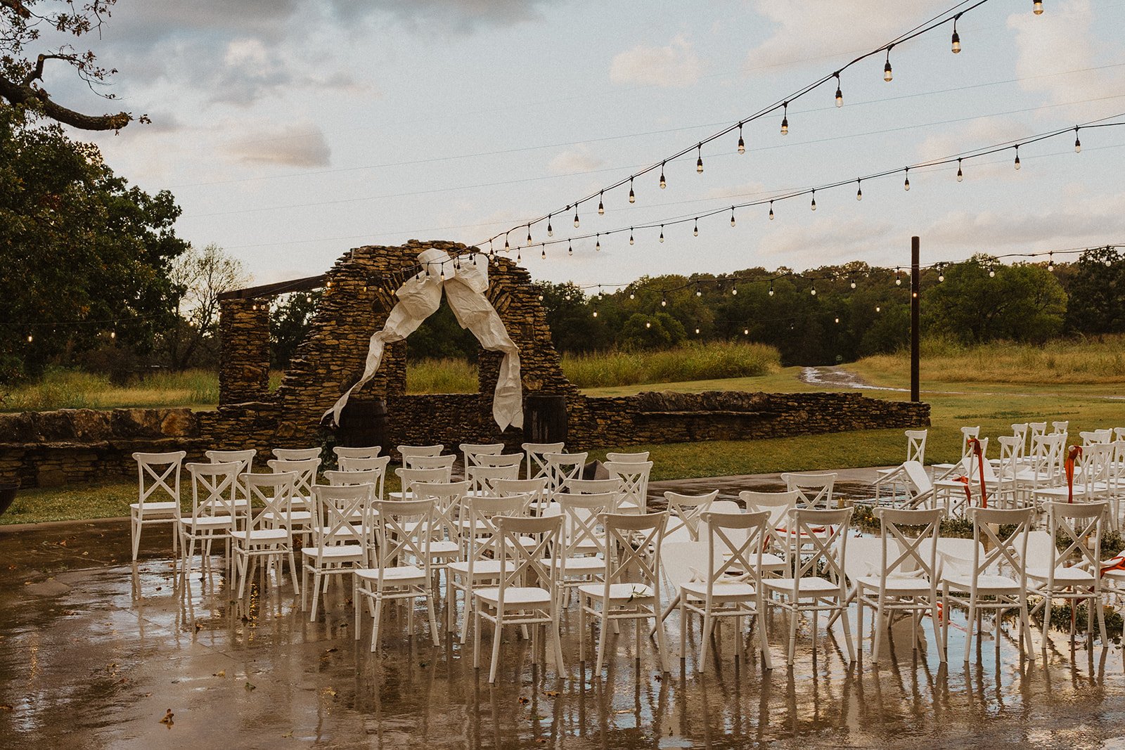 Rain-covered outdoor ceremony space at Bright Morning Farm with white chairs, string lights and a stone arch decorated with a large white bow.