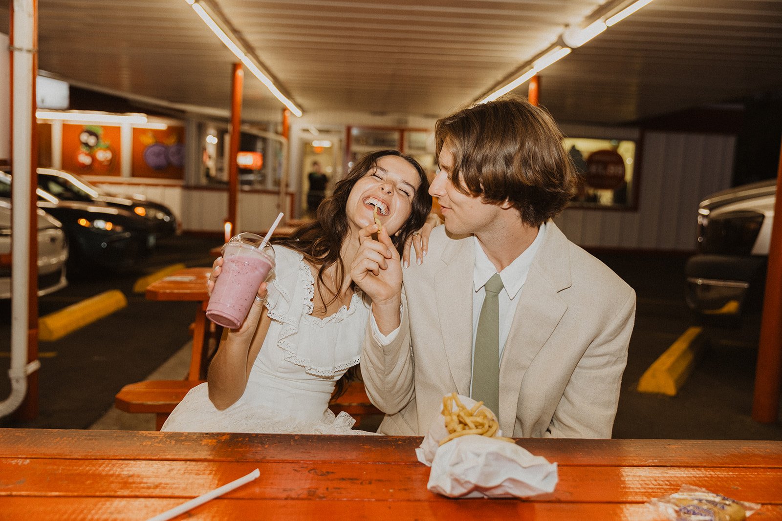 Bride and groom sharing fries and milkshake at retro drive-in after wedding