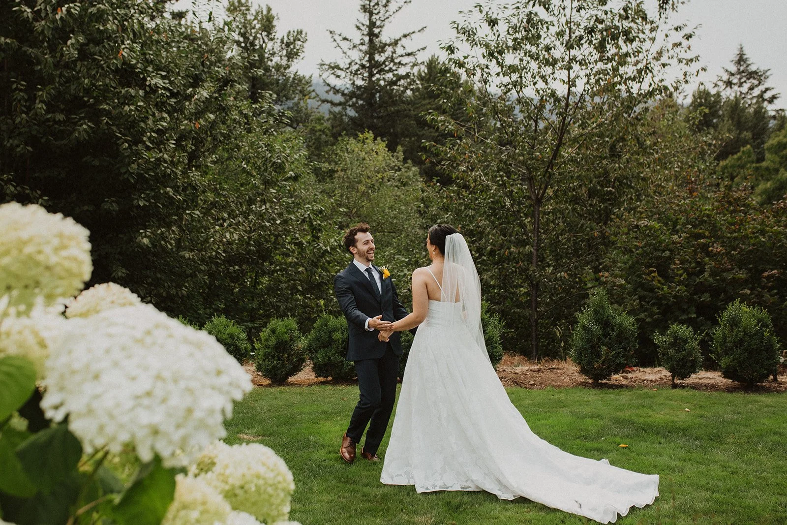 Bride and groom share a joyful first look in a lush garden wedding venue surrounded by greenery and white hydrangeas.