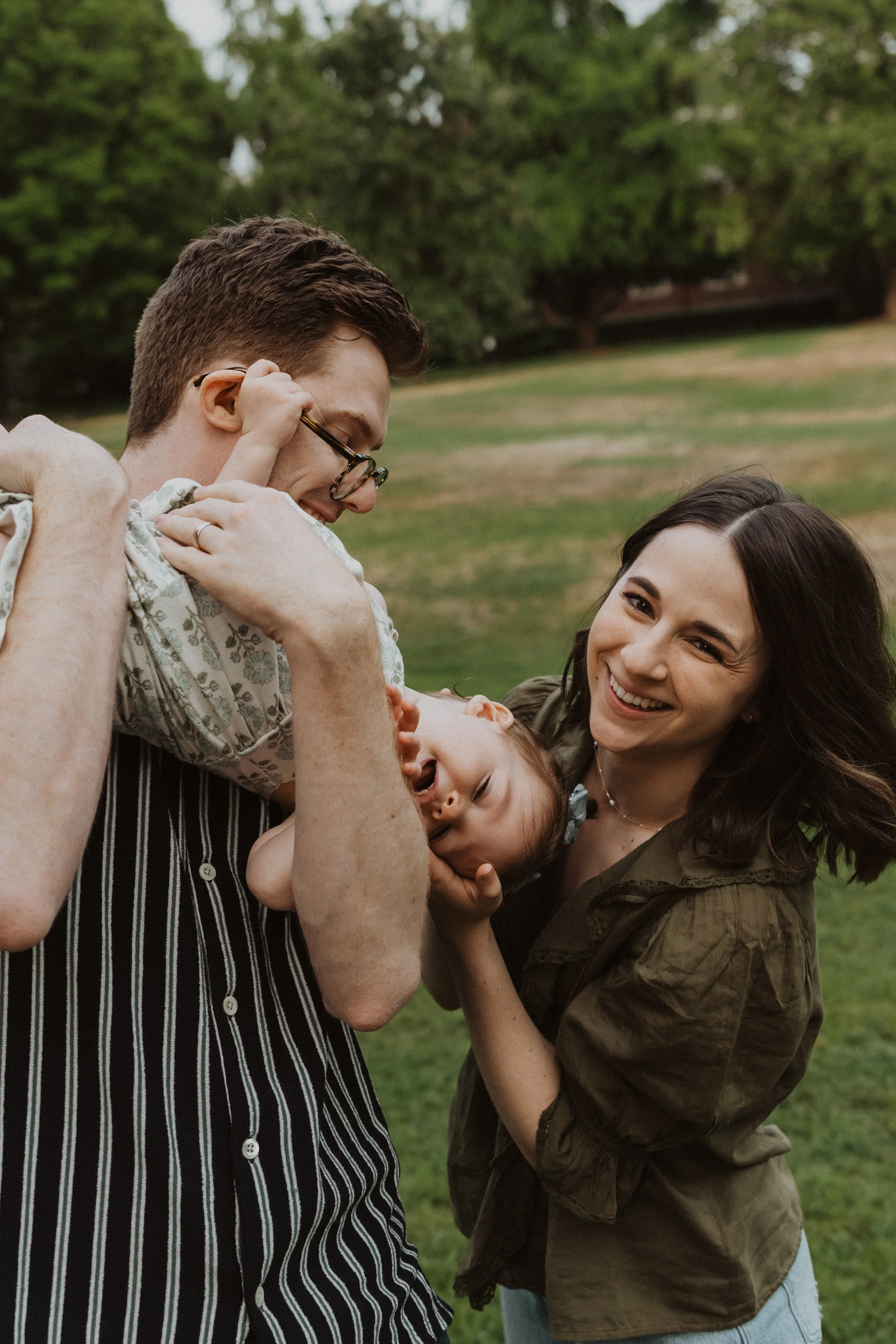 Family Session in Portland, Oregon. A happy family enjoying a moment outdoors, with a man and woman playing with a young girl, all smiling and laughing in a park.