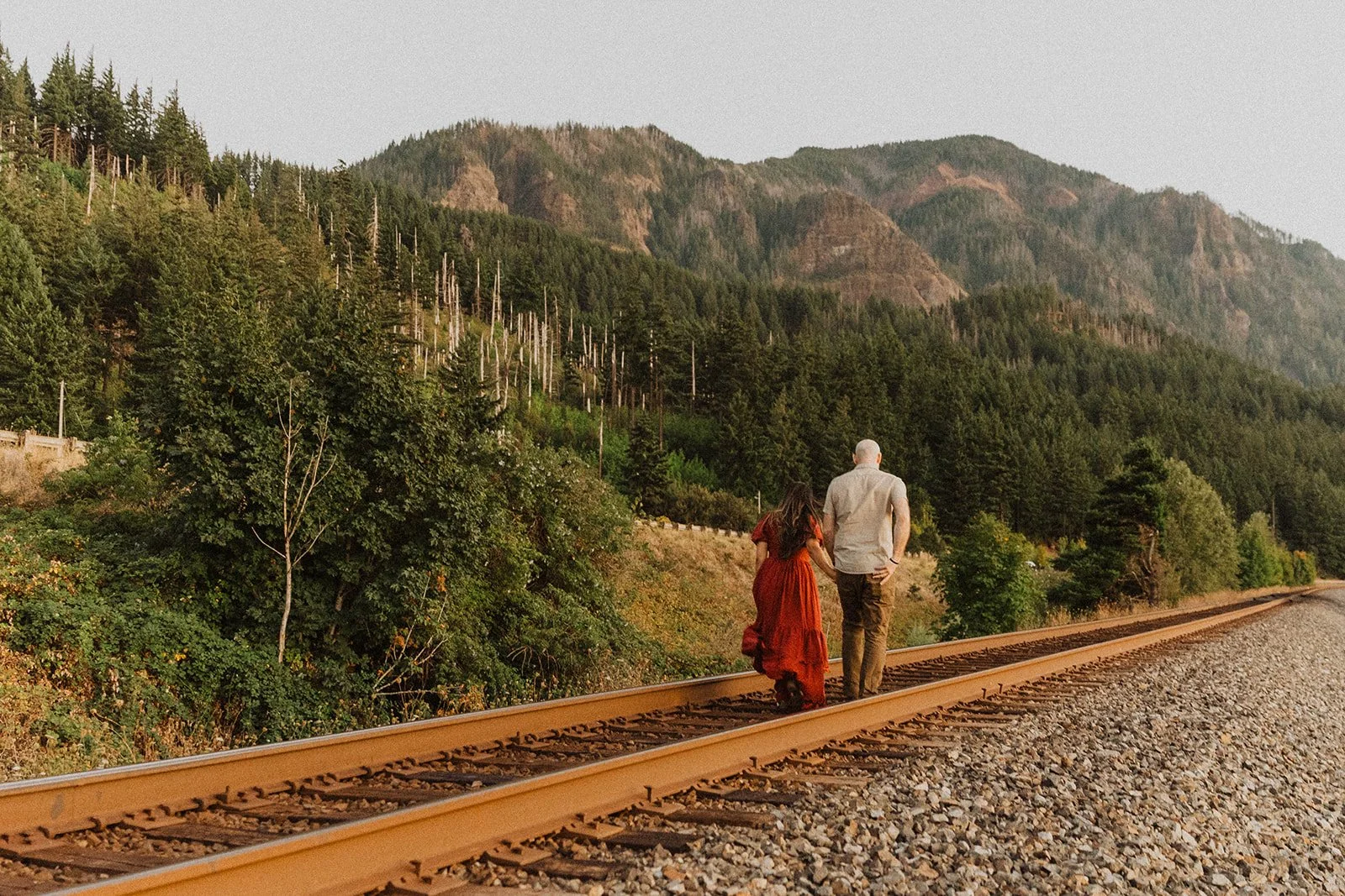 Couple walking hand in hand along railroad tracks surrounded by forested mountains during an adventurous outdoor engagement session at golden hour.
