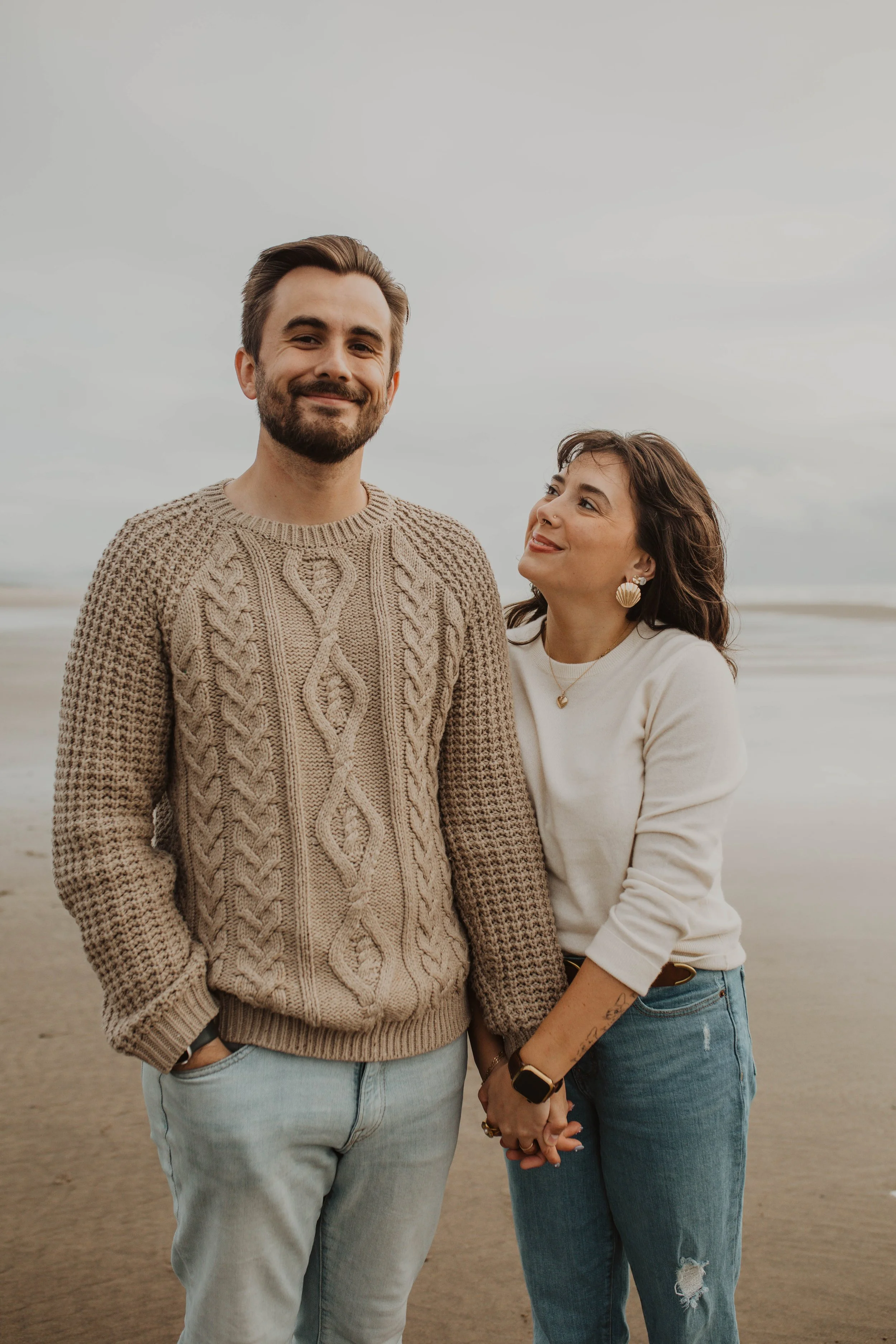 A couple standing on a beach, holding hands and smiling, with overcast skies in the background.