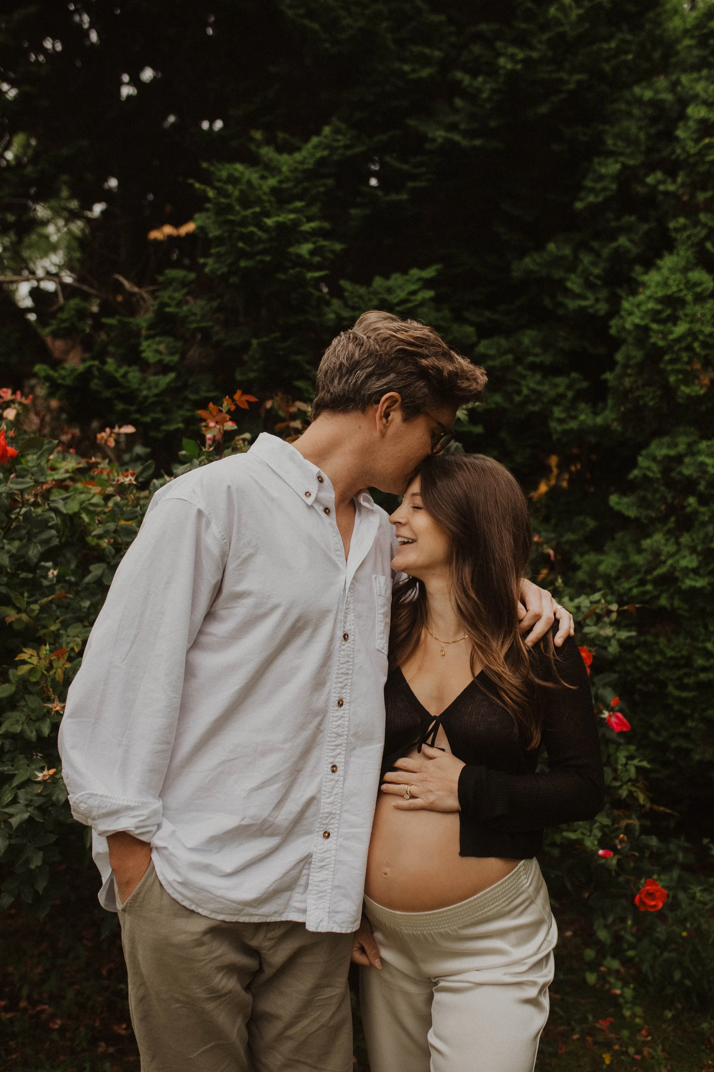 A couple standing close together outdoors, sharing a tender moment. The man is touching his forehead to the woman's forehead, and she is smiling with her eyes closed, holding her pregnant belly. They're surrounded by greenery and red flowers.