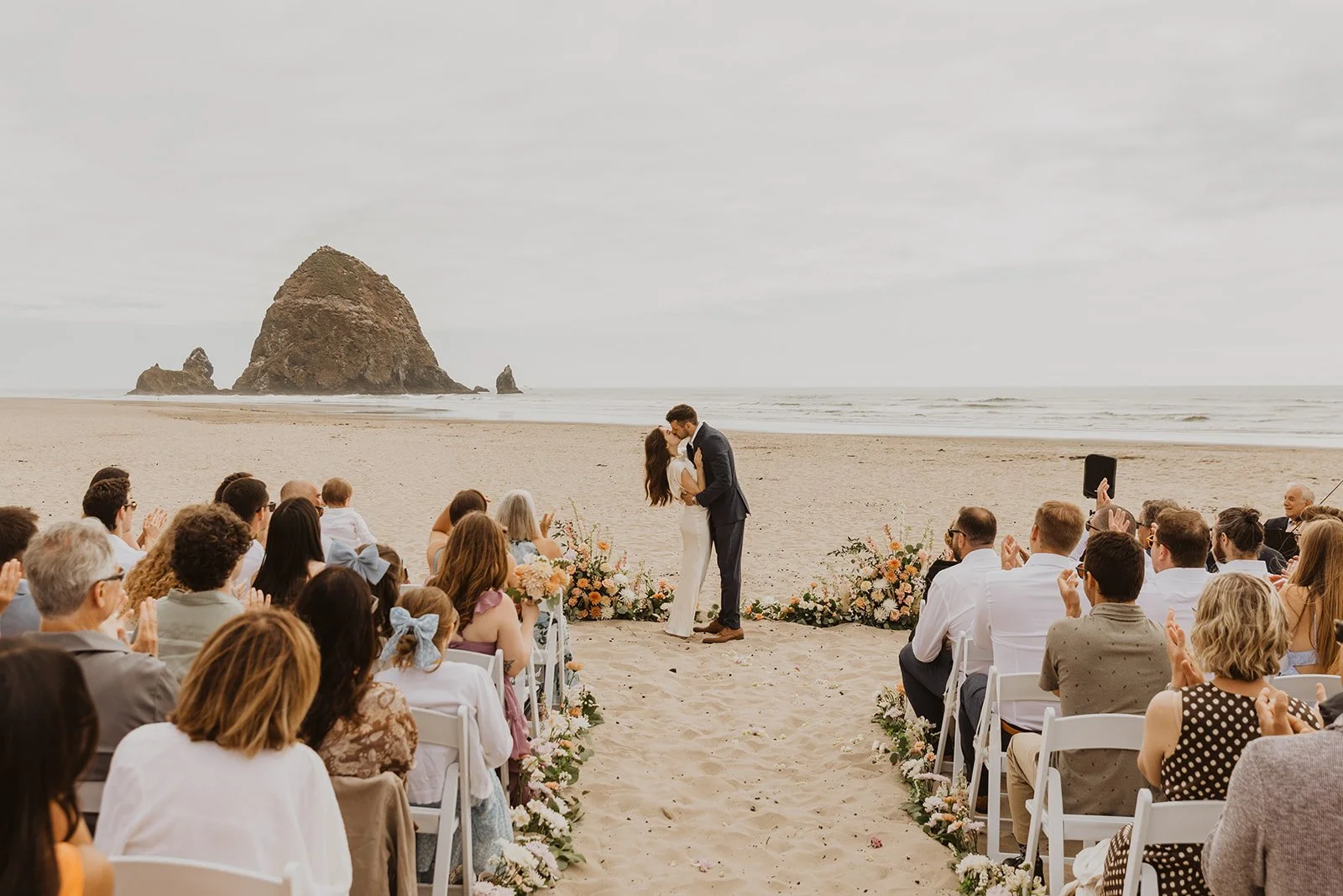 Couple sharing their first kiss during a beachfront wedding ceremony at Cannon Beach with guests celebrating around them