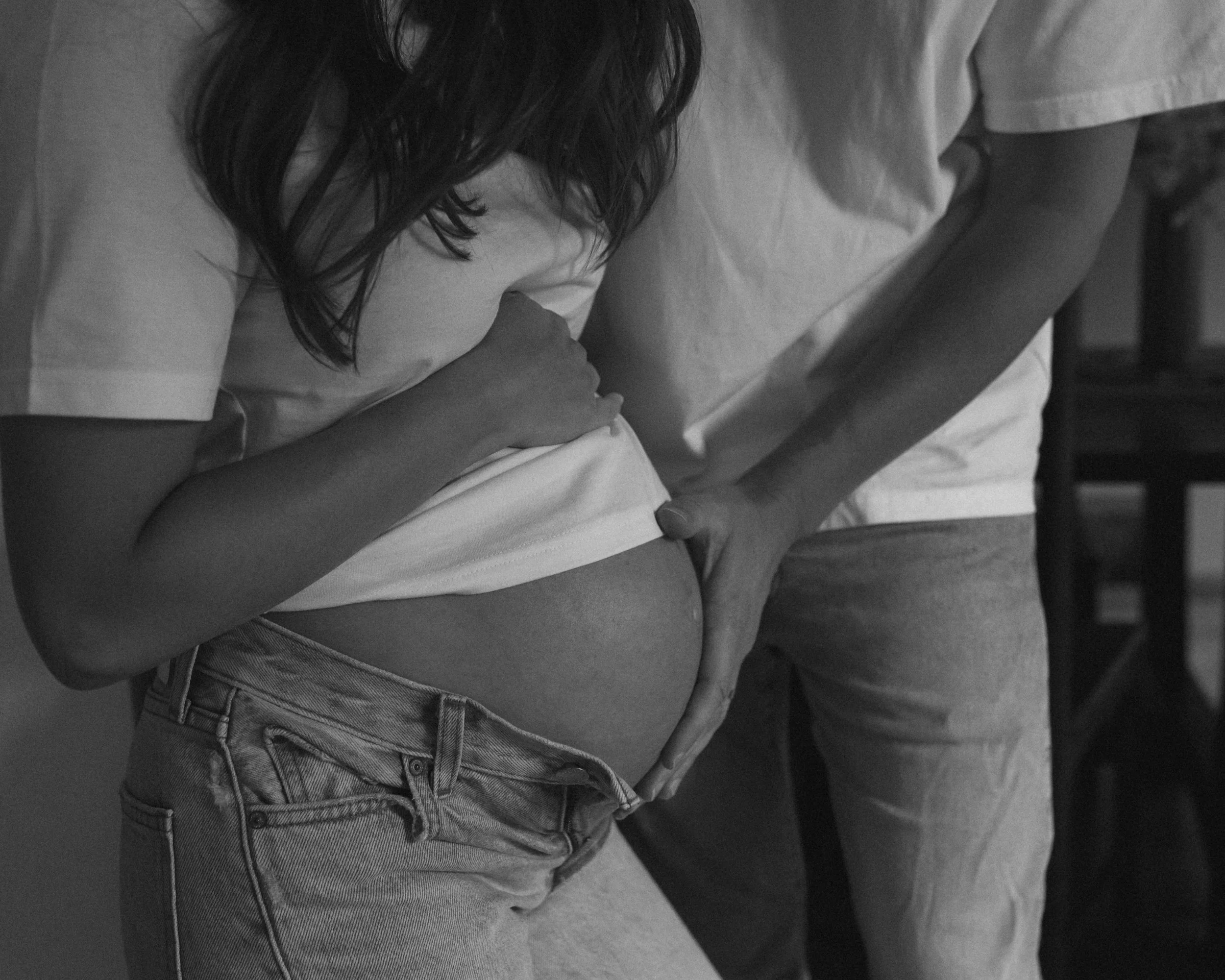 A woman and a man, both wearing casual clothing, are shown in a close-up black and white photo. The woman is holding her shirt up to reveal her lower abdomen, with her hand on her stomach. The man is standing beside her, with his hand on her waist, gently tracing her side.