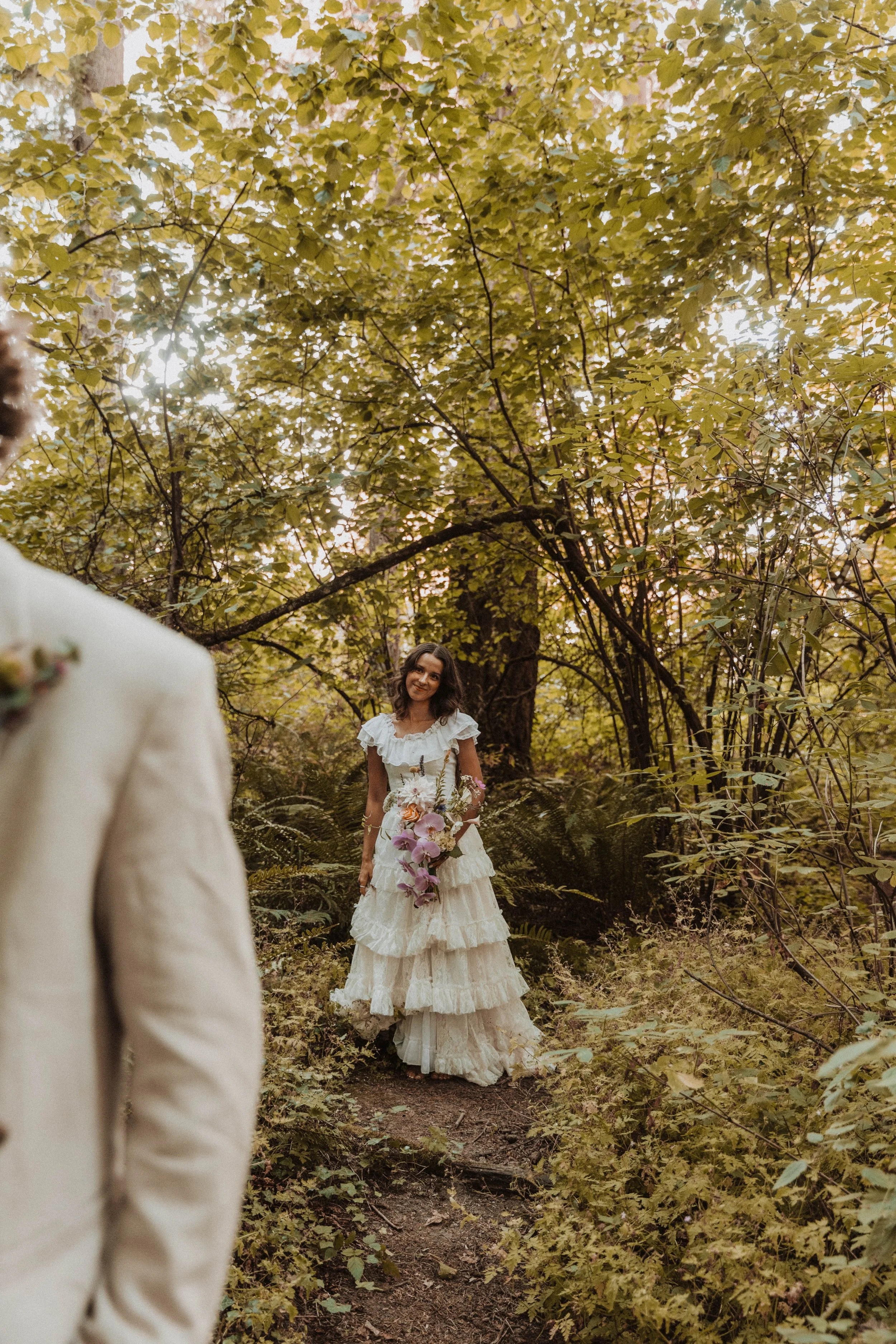 Bride standing in a forest clearing holding a colorful bouquet while her partner approaches, documented by a wedding photographer in Portland Oregon.