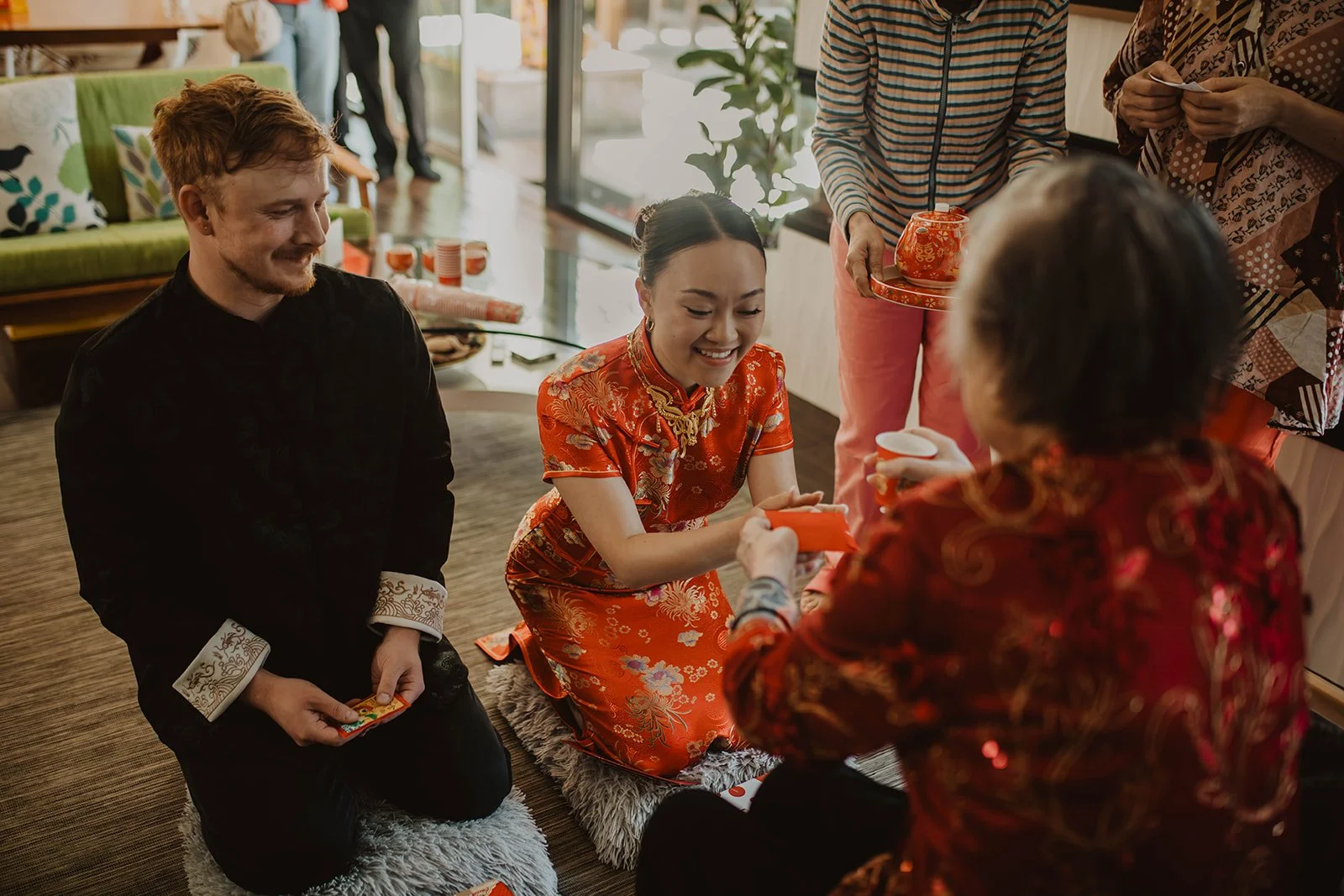 Bride and groom kneel during a Chinese tea ceremony as an elder presents a red envelope and wedding blessings.