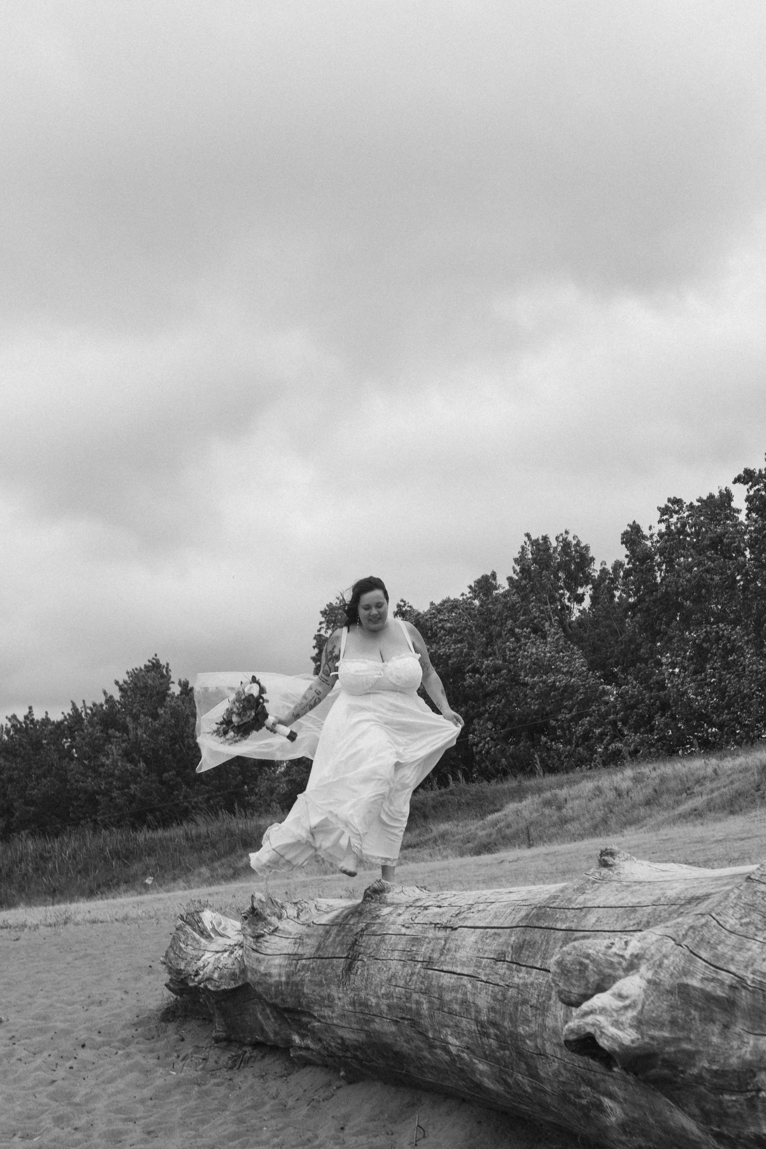 Bride in a flowing dress running across a fallen log in a grassy field, captured in a candid black and white moment by an Oregon wedding photographer.