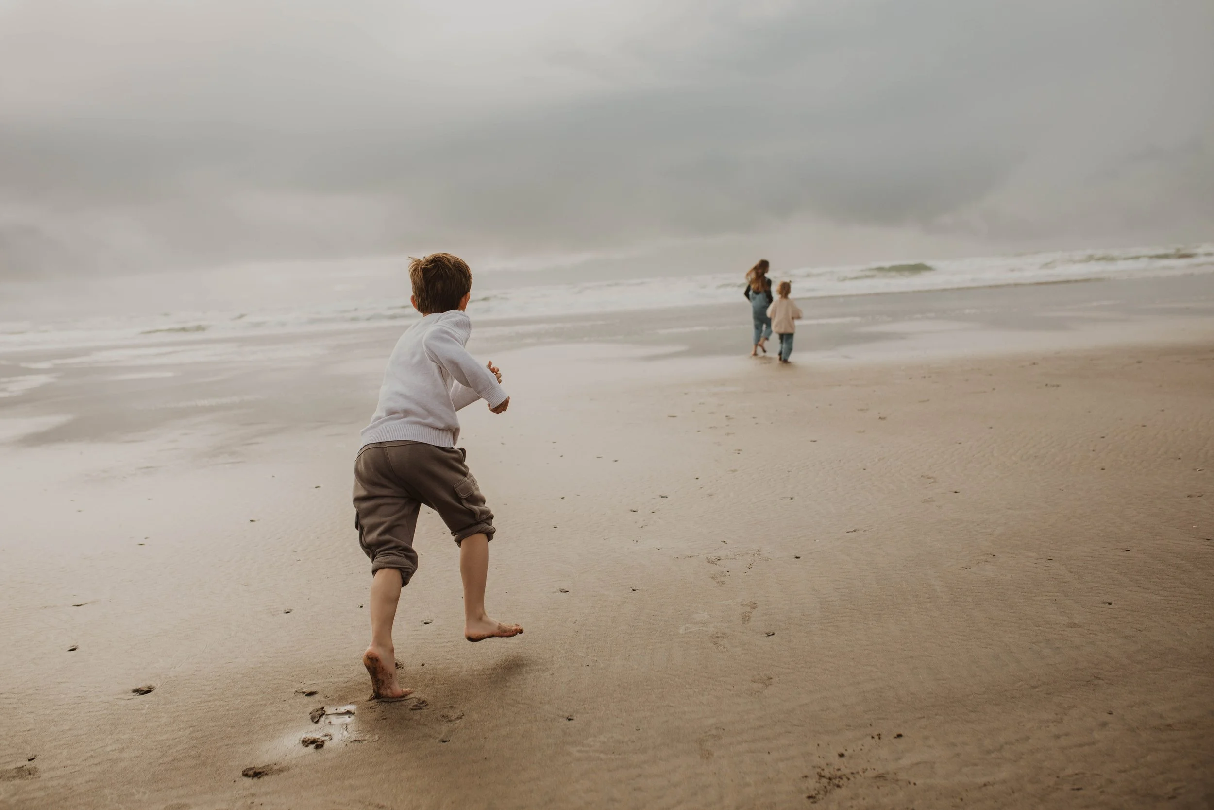 A young boy running on the beach chasing two women, with an overcast sky and waves in the background.