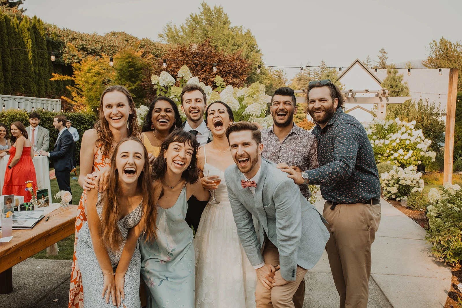 Group of wedding guests and friends laugh together while posing for a fun outdoor photo during a garden wedding reception with string lights and hydrangeas in the background.