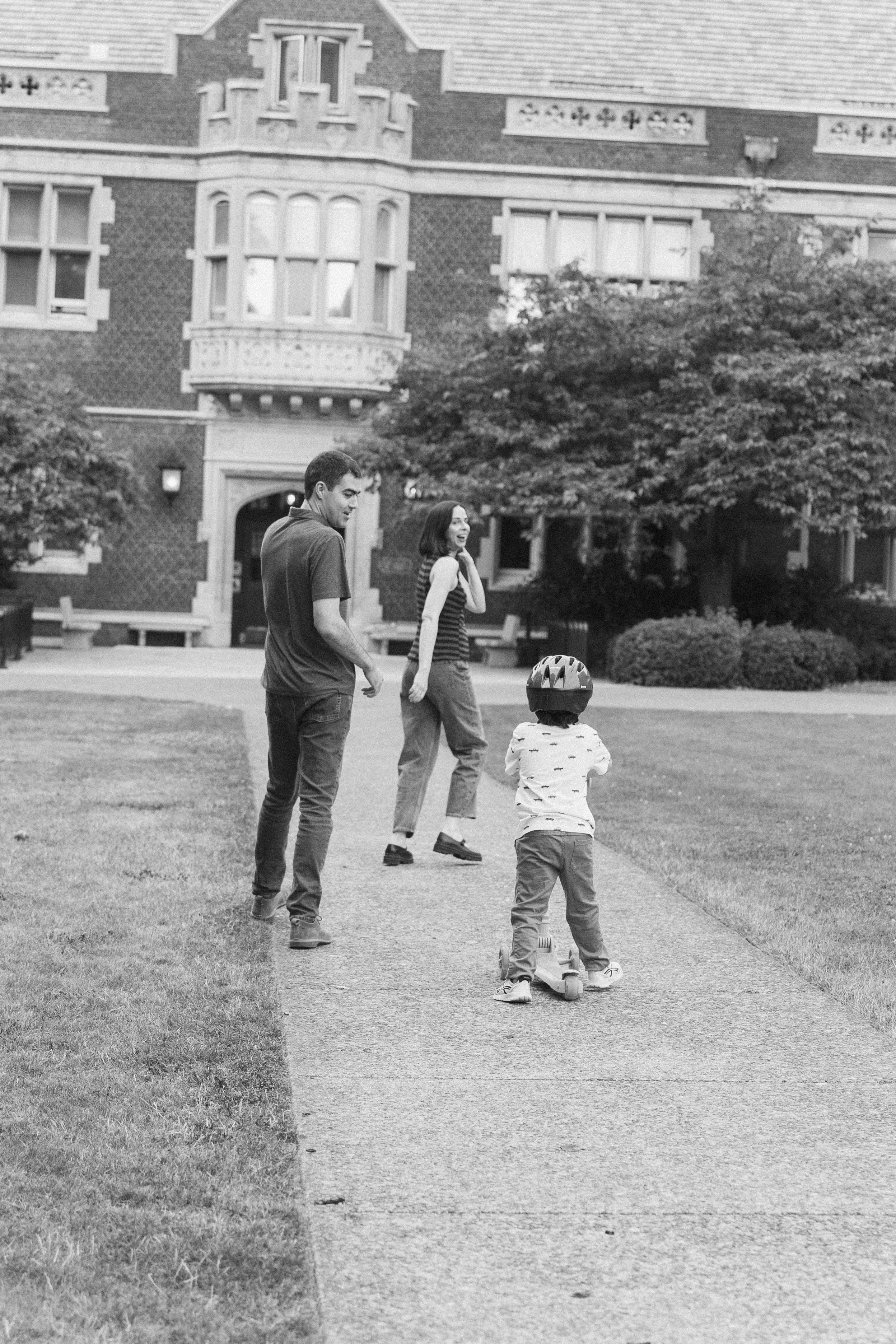 A family of three, including a woman, man, and young boy with a helmet, walking on a sidewalk in front of a large brick house with trees and bushes.