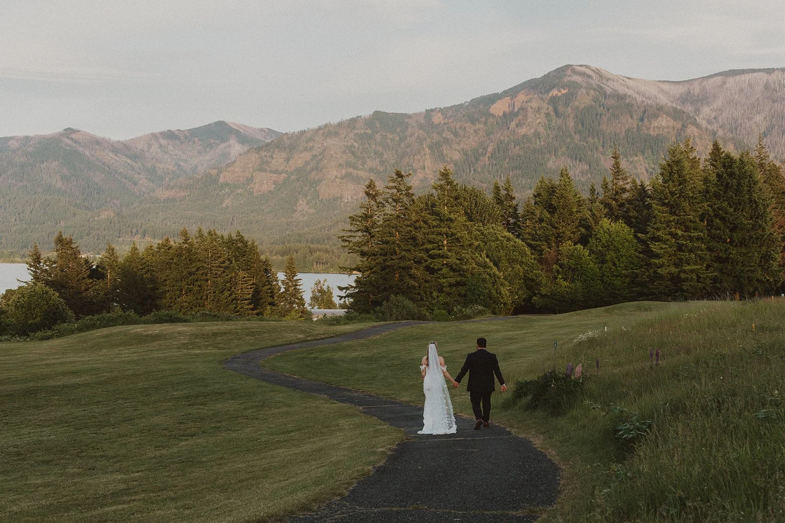 Newlyweds walking hand in hand along a winding path at Skamania Lodge with Columbia River Gorge views in the background.