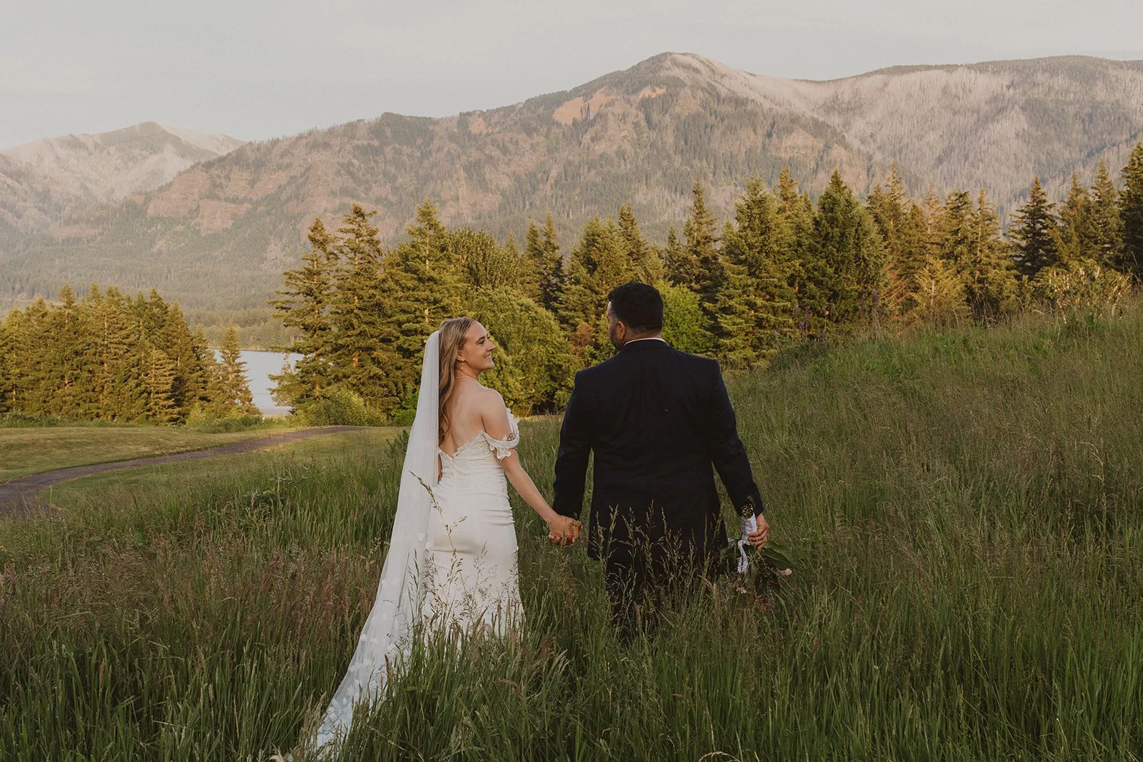 Bride and groom walking together through tall grass with mountain views surrounding Skamania Lodge.