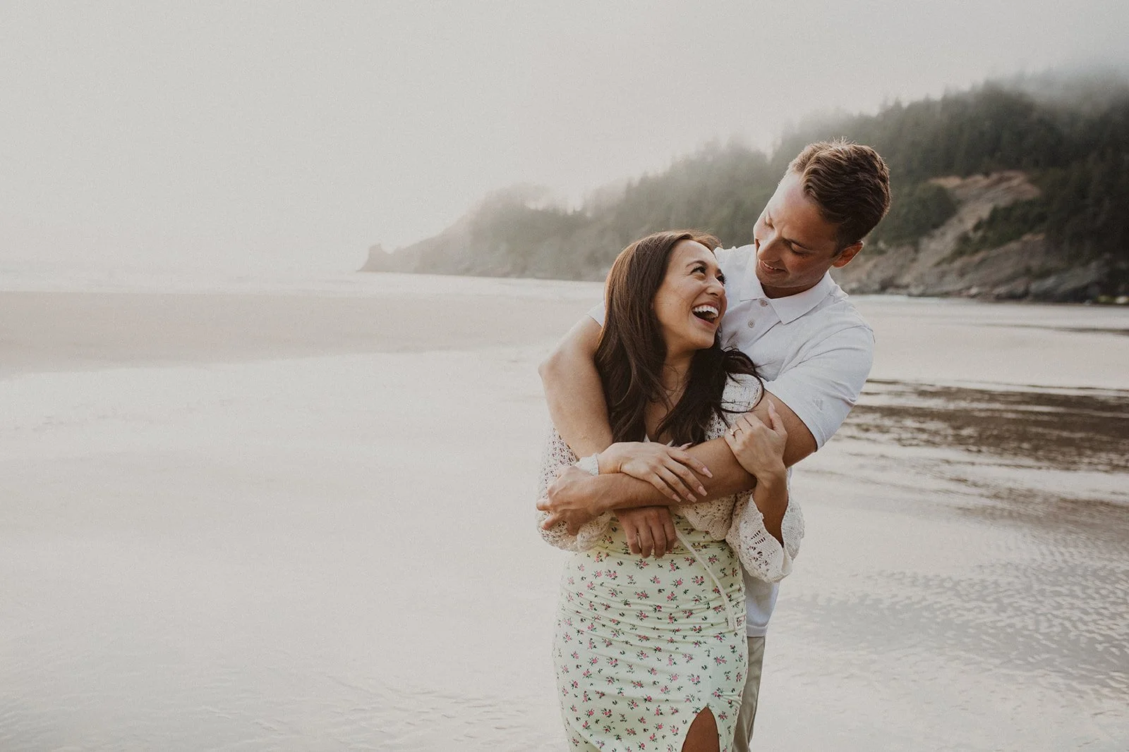 Couple laughing and embracing on the beach during a romantic Oregon coast engagement session with soft coastal fog