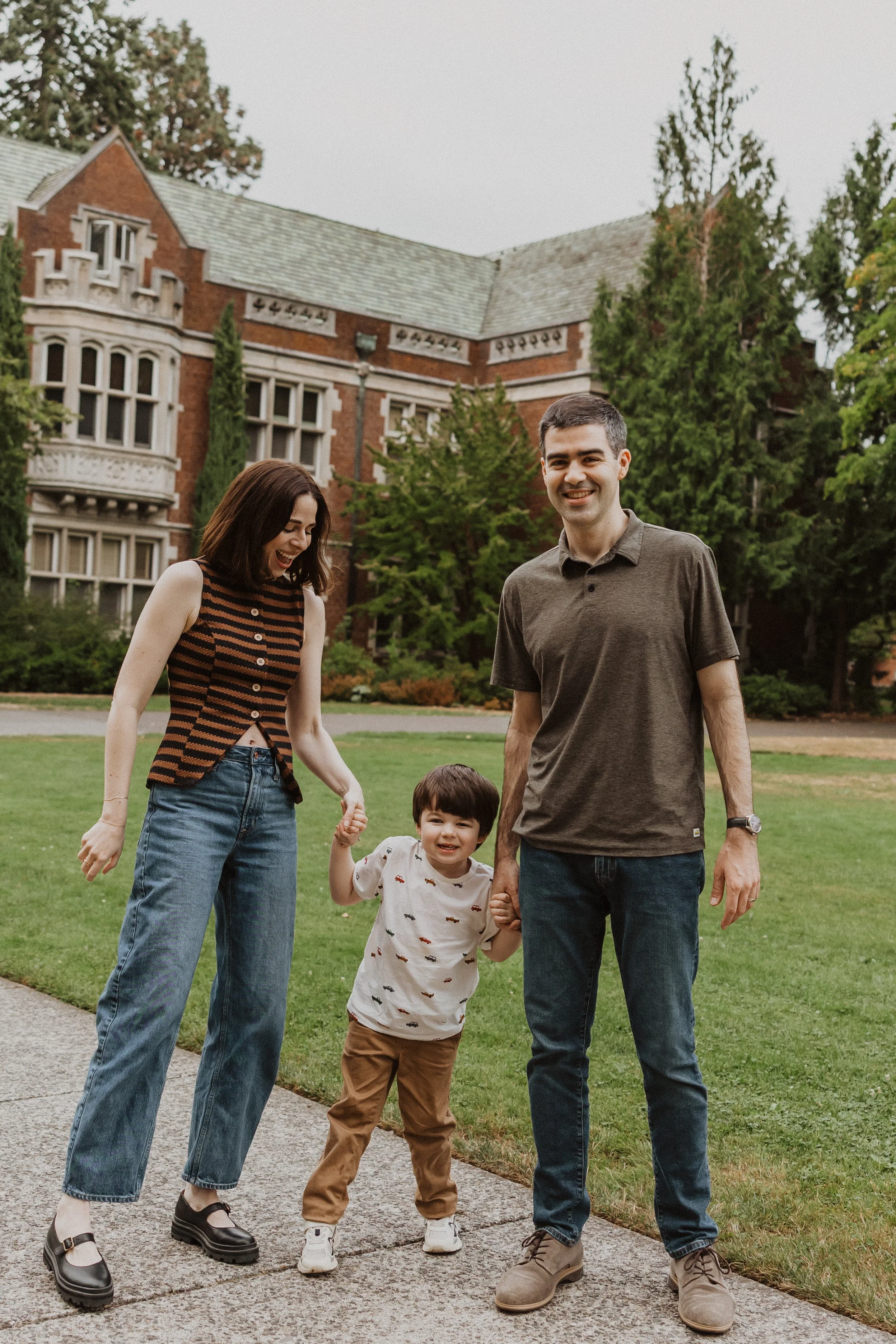 A family of three holding hands and smiling on a sidewalk in front of a large, old brick house surrounded by trees.