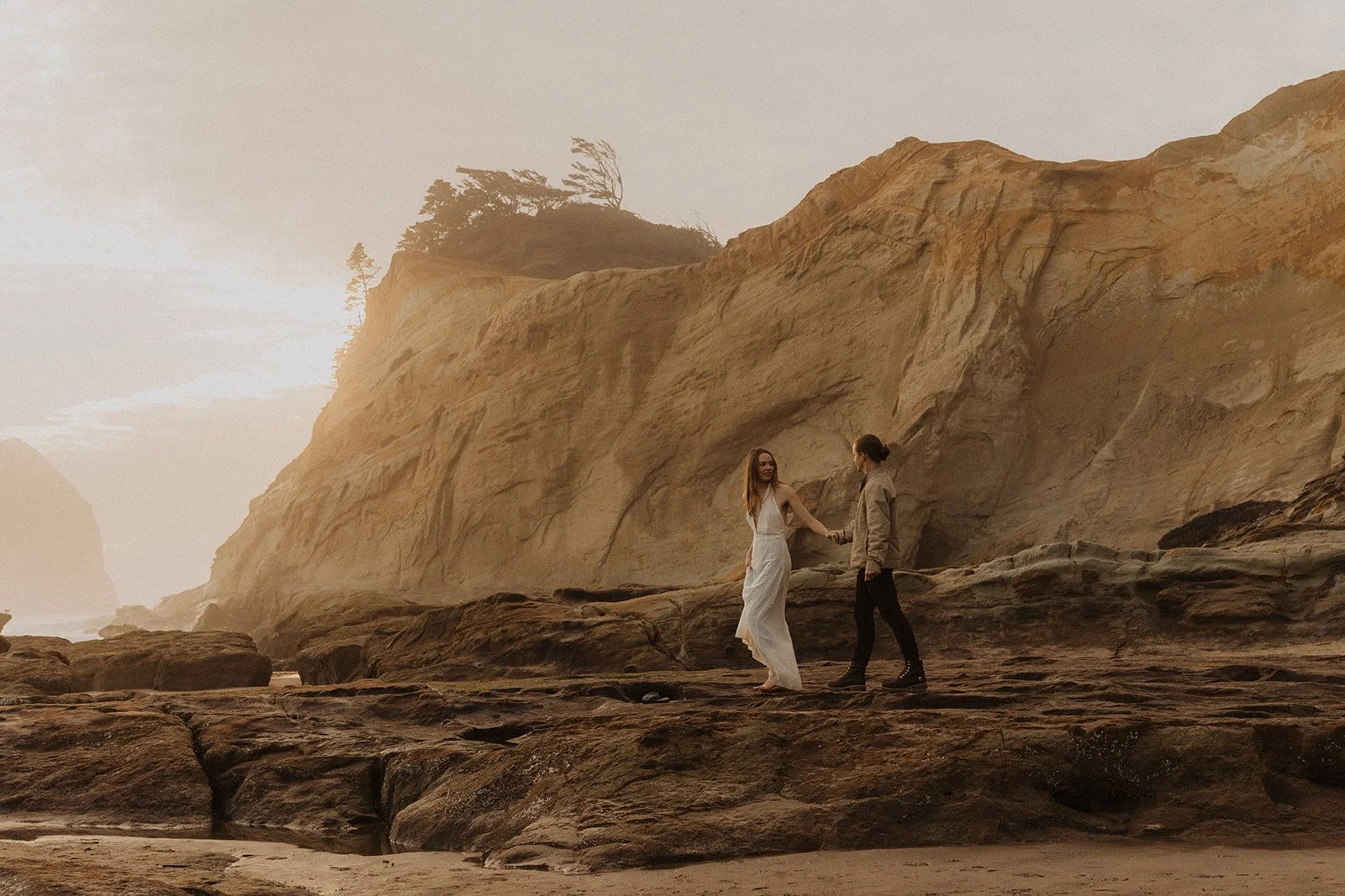 Couple walk hand in hand across the rocky shoreline beneath the sandstone cliffs at Cape Kiwanda during their engagement session