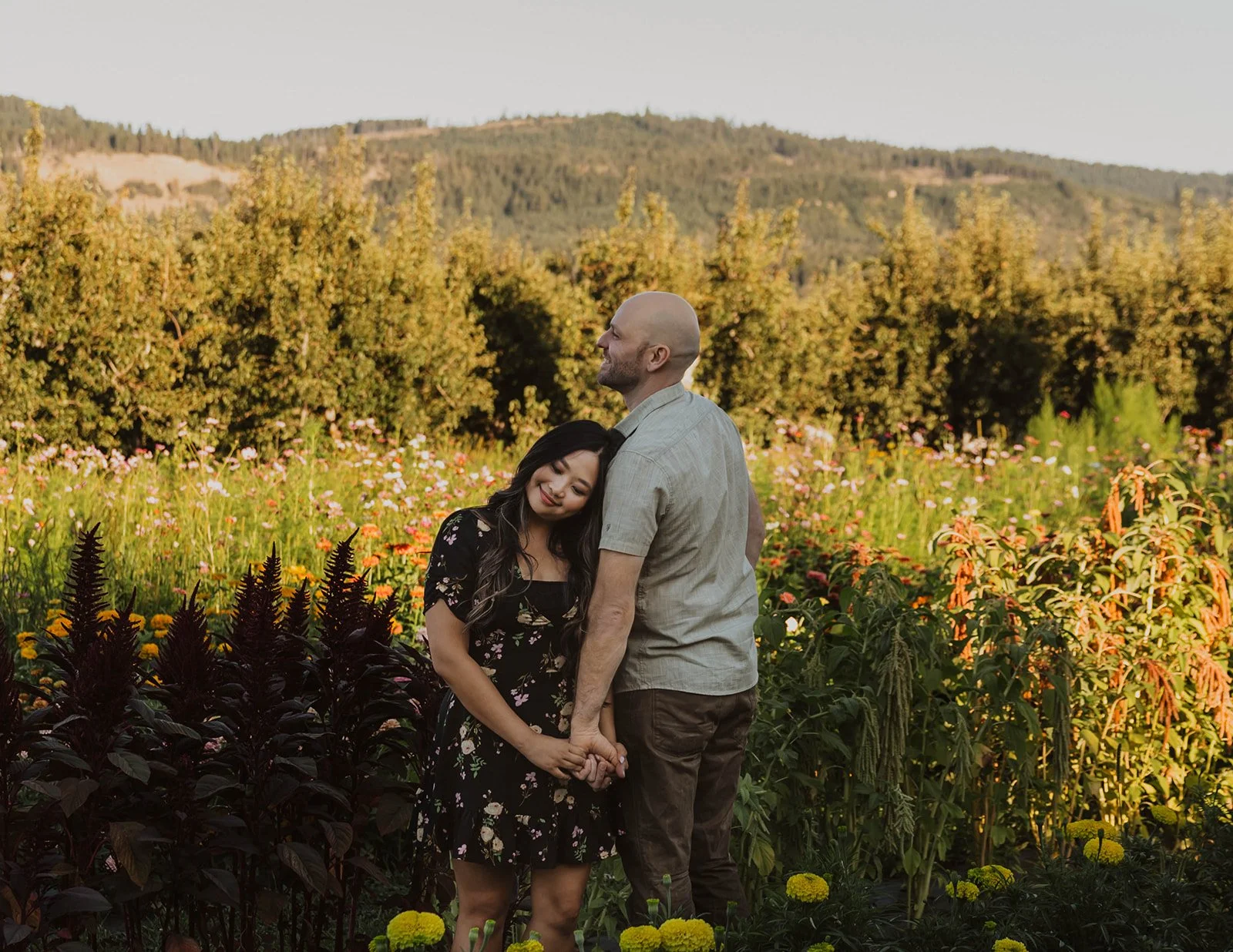 Engaged couple holding hands in a flower garden with rolling hills and orchard views in the background during golden hour.
