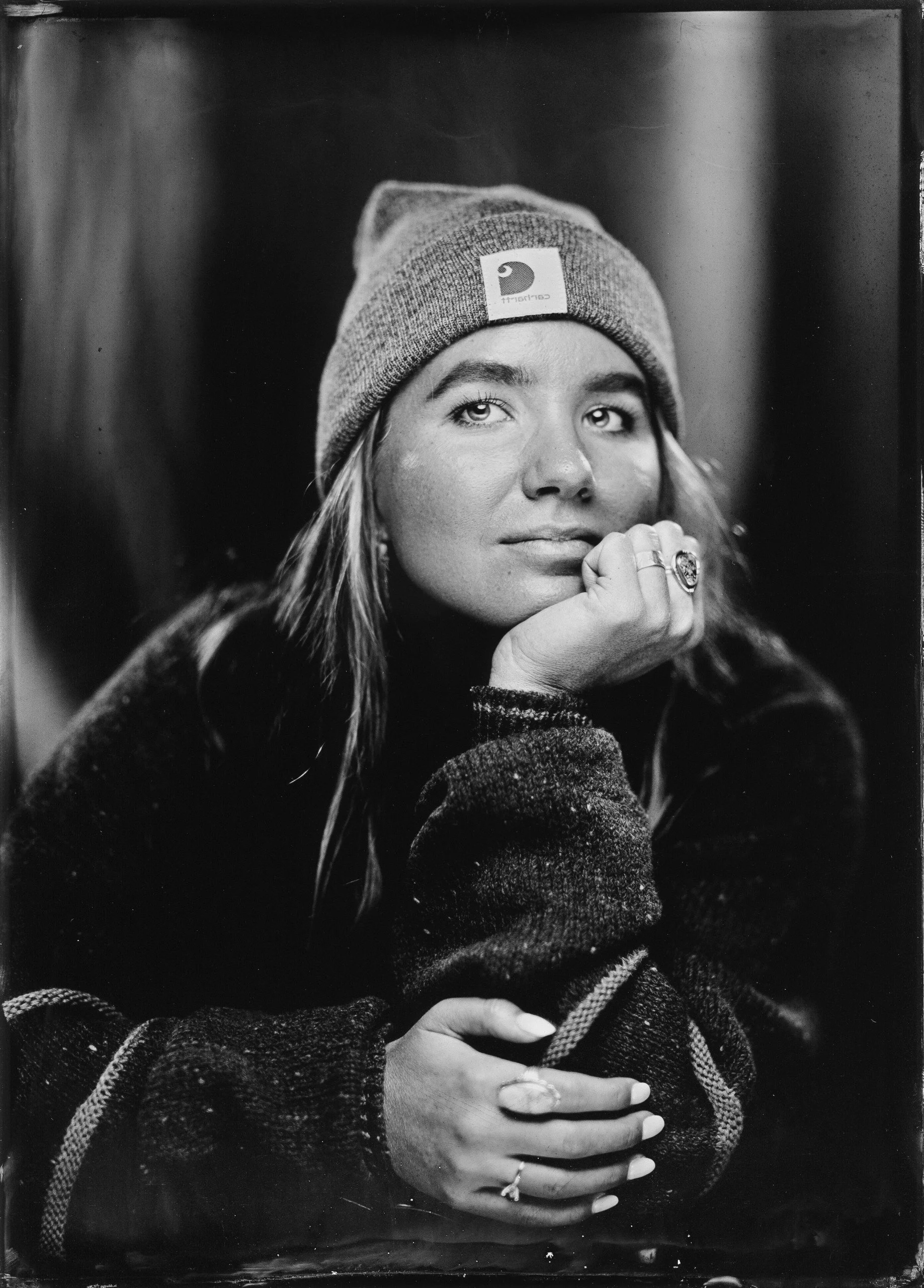Black and white portrait of an  Oregon wedding photographerwearing a beanie hat, resting her chin on her hand and looking at the camera. She has long hair, rings on her fingers, and is dressed in a sweater.