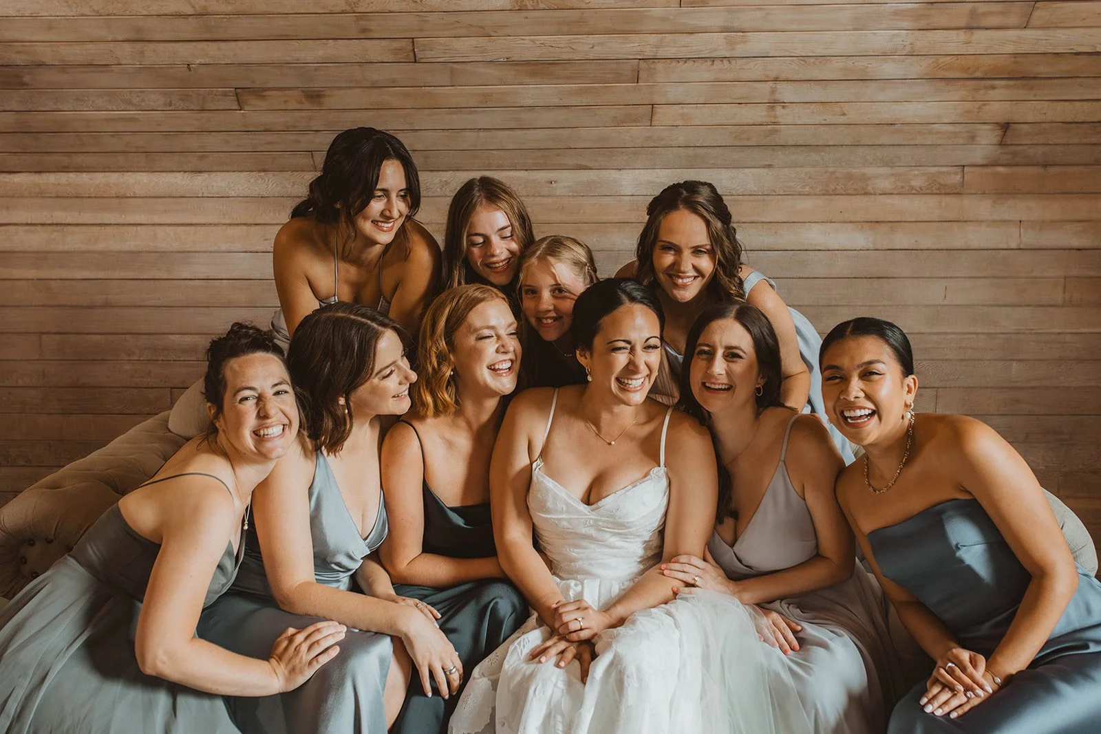 Bride laughs while surrounded by bridesmaids and flower girls in blue dresses during a joyful getting-ready group portrait at Cape Horn Estate