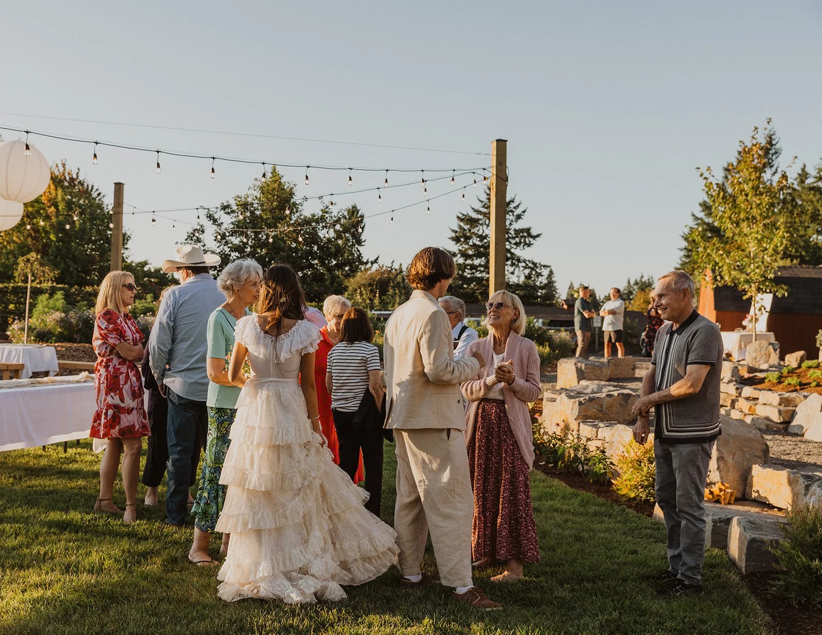 Wedding guests mingling on a grassy outdoor reception space at sunset, with the bride in a tiered lace dress and the groom in a light suit chatting among friends under string lights
