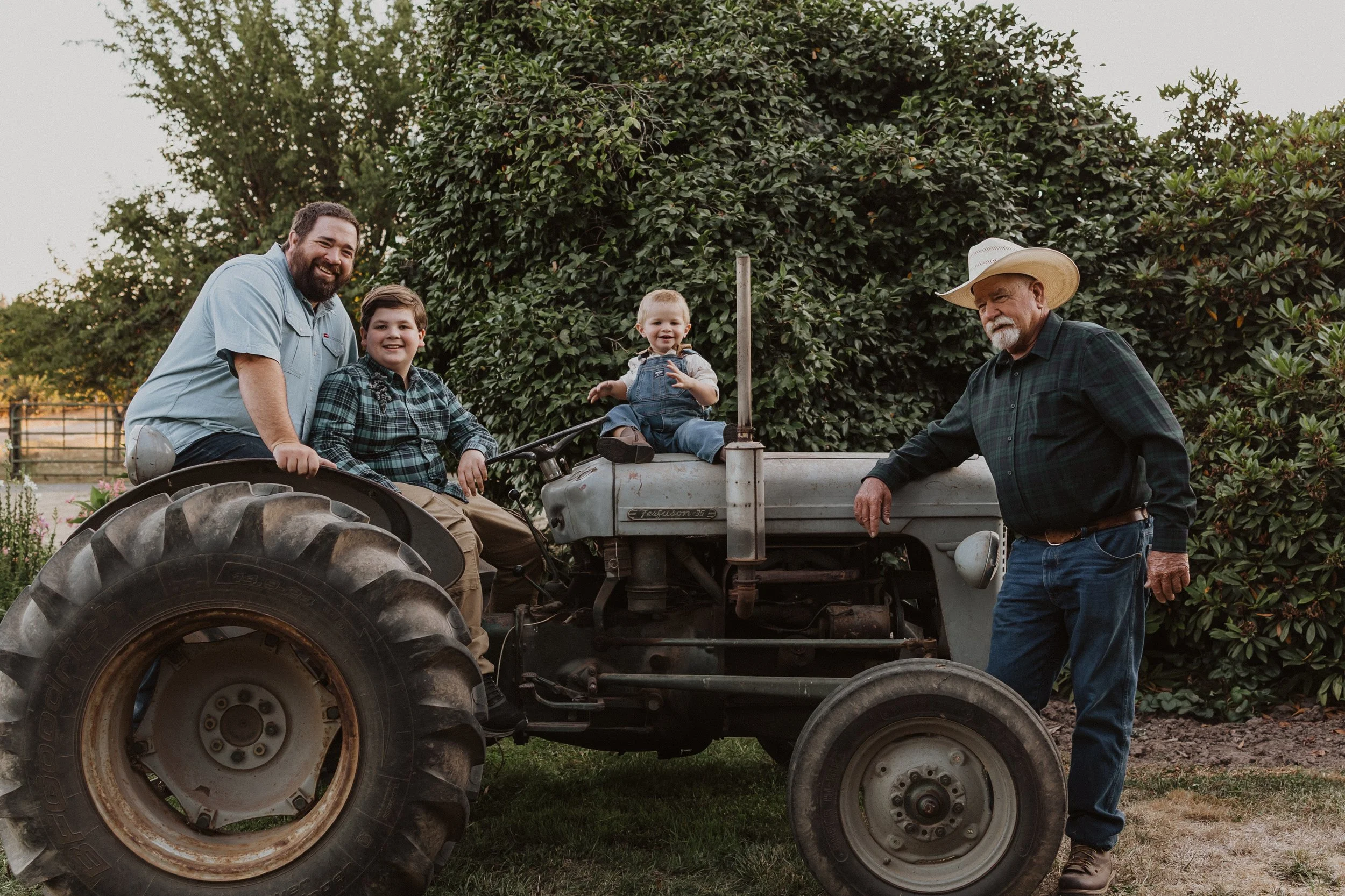 Family Session in Aurora, Oregon. Three generations of men on a vintage tractor outdoors, with trees and bushes in the background. An older man in a cowboy hat stands next to the tractor, while a man and two children sit on the tractor, smiling.