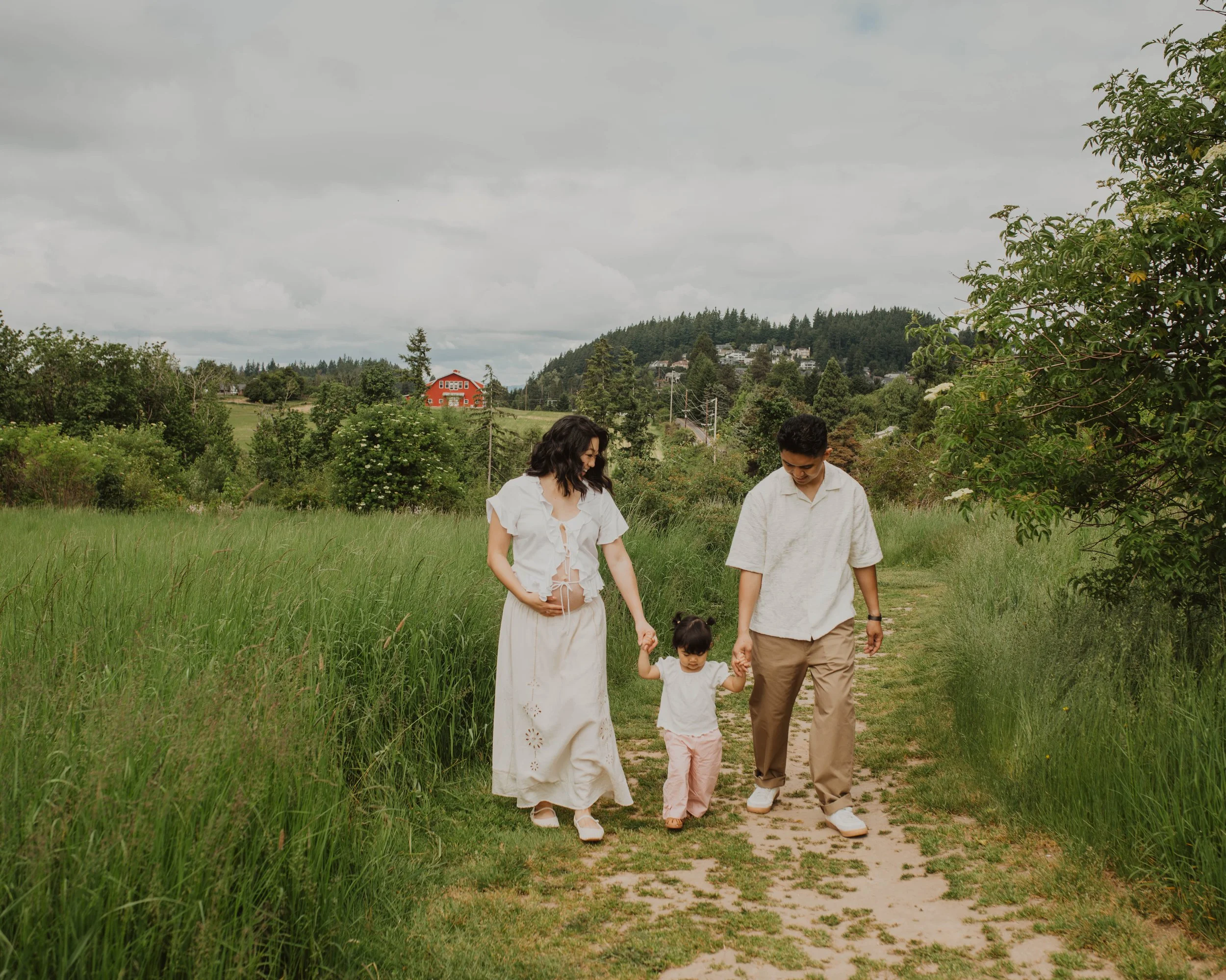 A family of three walking on a dirt path through a grassy field, holding hands and smiling, with a woman on the left, a man on the right, and a young girl in the middle.