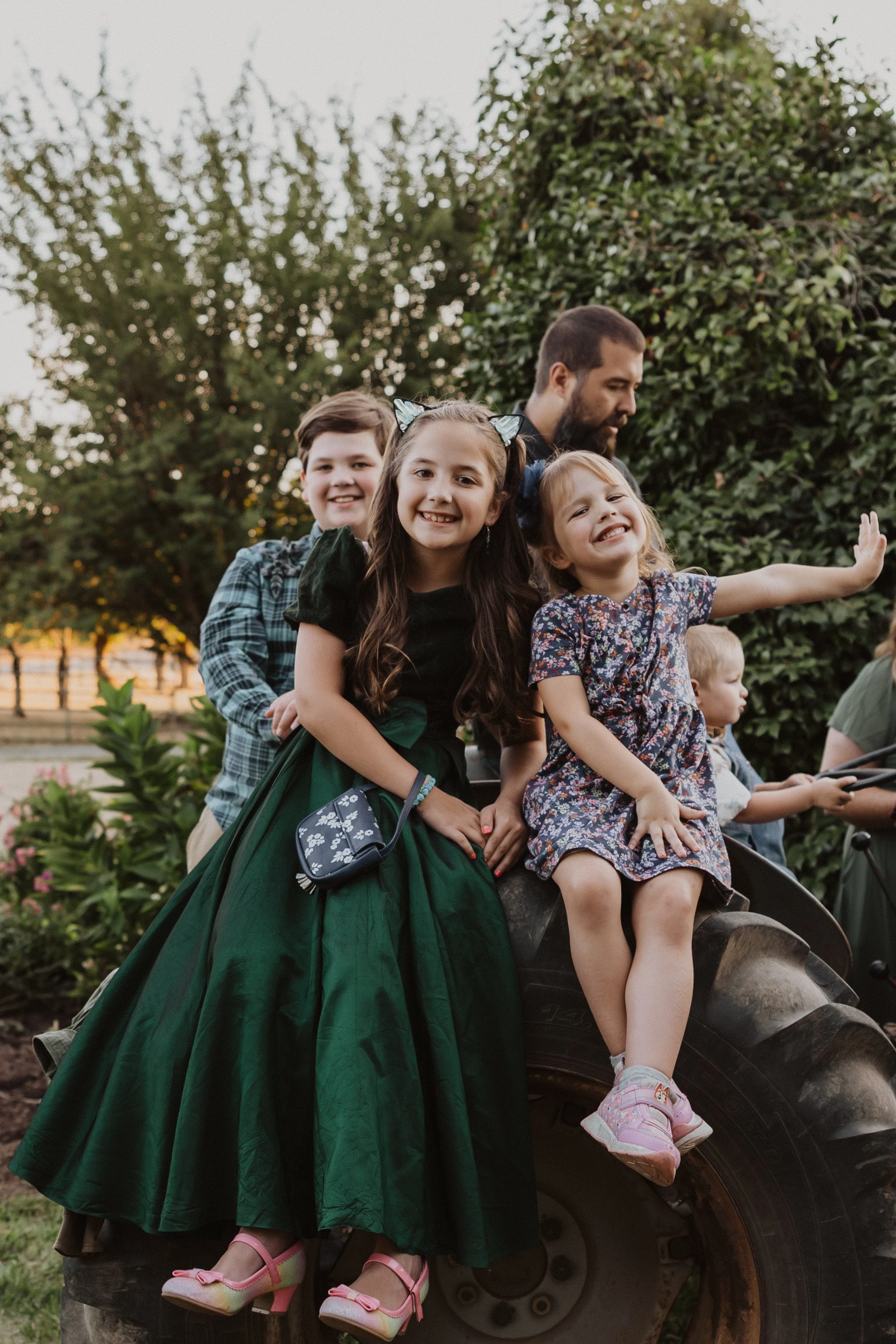 Family Session in Aurora, Oregon on the Koch Family Farm. A group of children and an adult sitting on a tractor tire outdoors, smiling and waving.