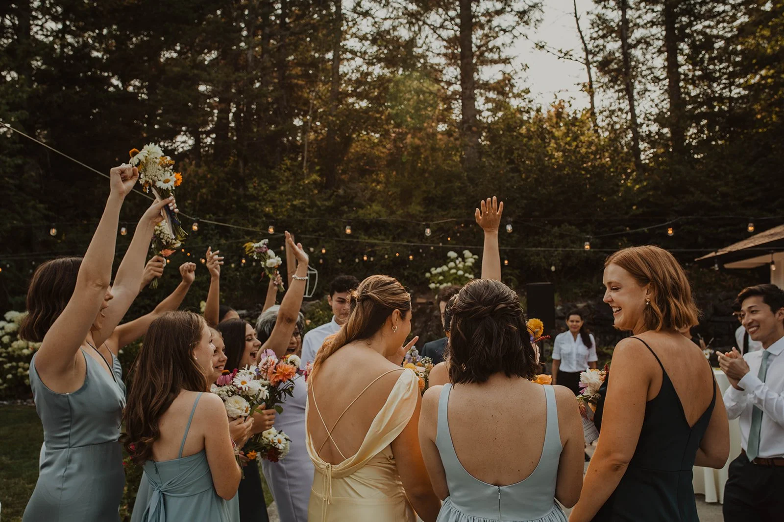 Wedding guests and bridal party members cheering and raising bouquets during an outdoor reception at sunset under string lights surrounded by trees.