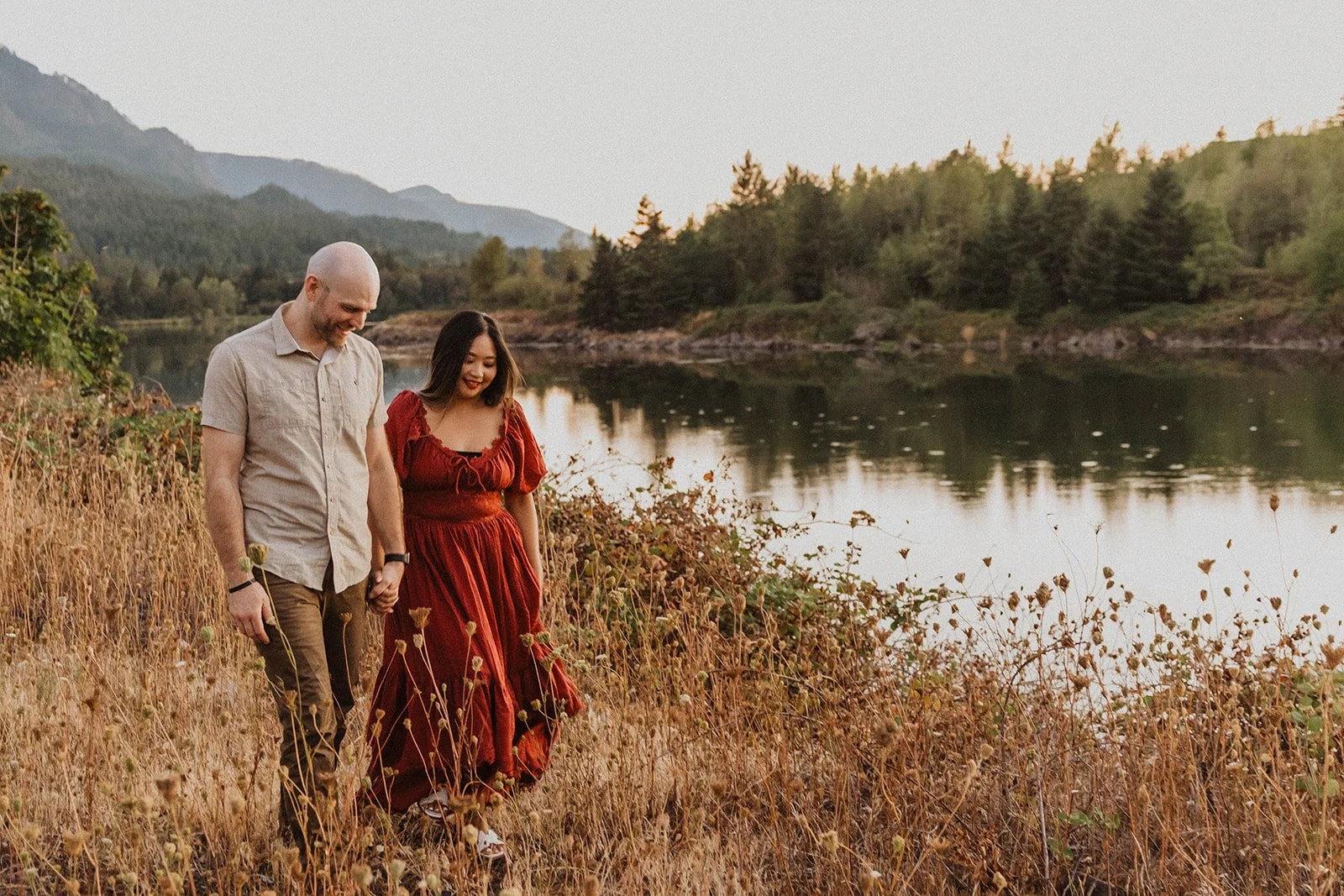 Couple holding hands and walking through a grassy riverside landscape during their Mt Hood engagement photos