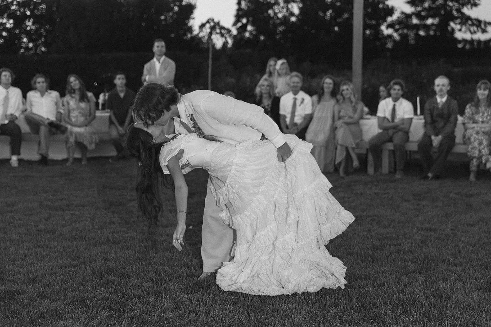 Black-and-white photo of the bride and groom sharing a kiss during their first dance, with the groom dipping the bride on a lawn as guests watch in the background