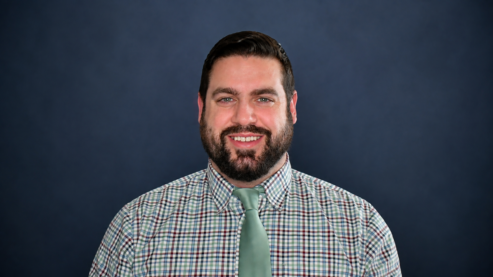 A smiling man with dark hair and beard, wearing a green sweater over a checkered shirt, against a plain, light-colored background.