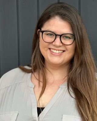 A woman with long brown hair, glasses, and a light-colored shirt smiling in front of a dark gray background.
