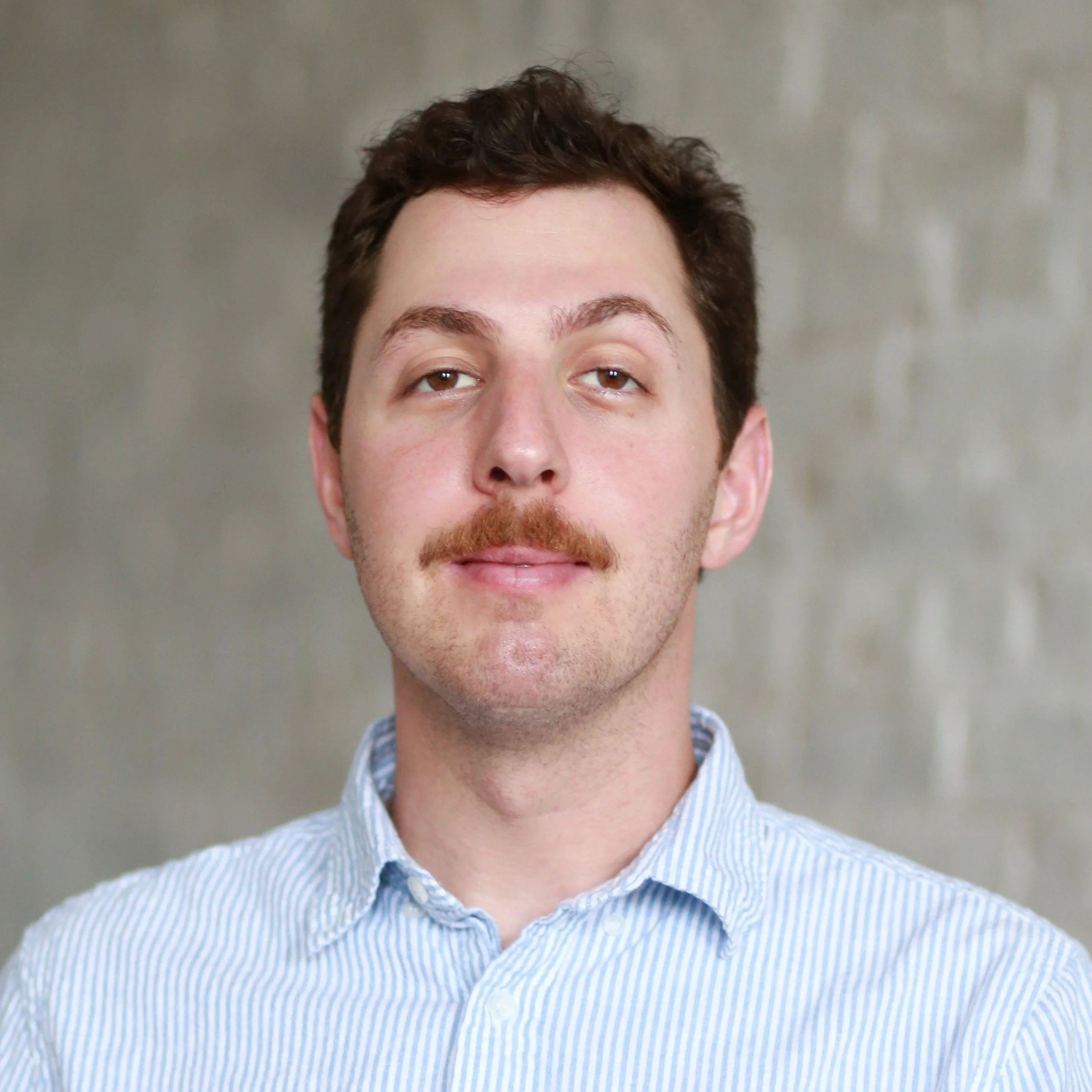 Close-up of a young man with a mustache and brown hair, wearing a light blue striped button-up shirt, against a blurred neutral background.