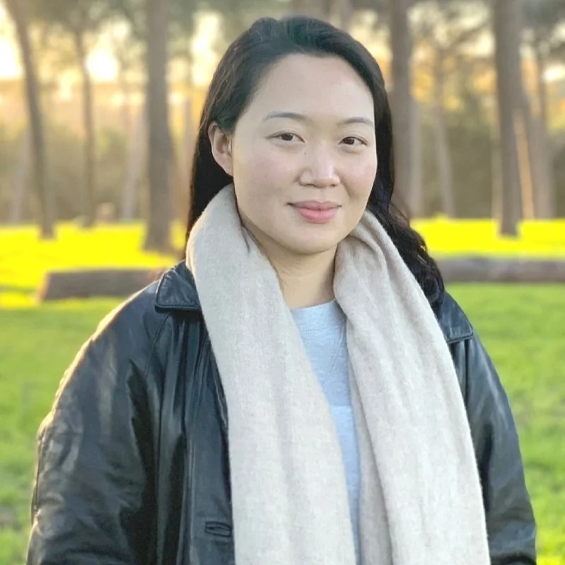 A woman with long dark hair, wearing a black leather jacket and a cream-colored scarf, standing outdoors in a park with trees and yellow benches in the background during golden hour.