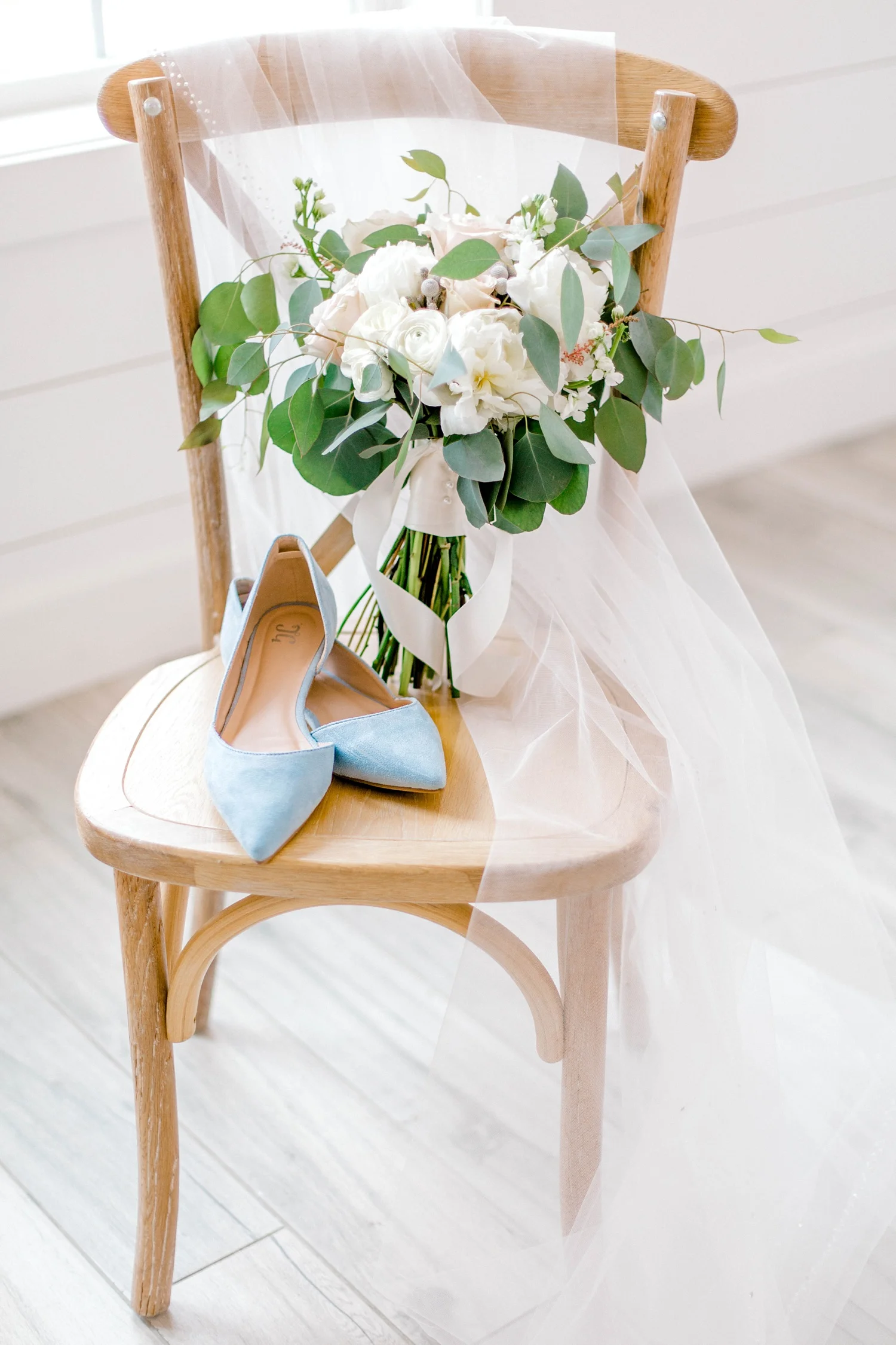 Bride’s wedding bouquet and blue shoes sitting on a chair with the bride’s veil. Eucalyptus and roses bridal wedding bouquet.