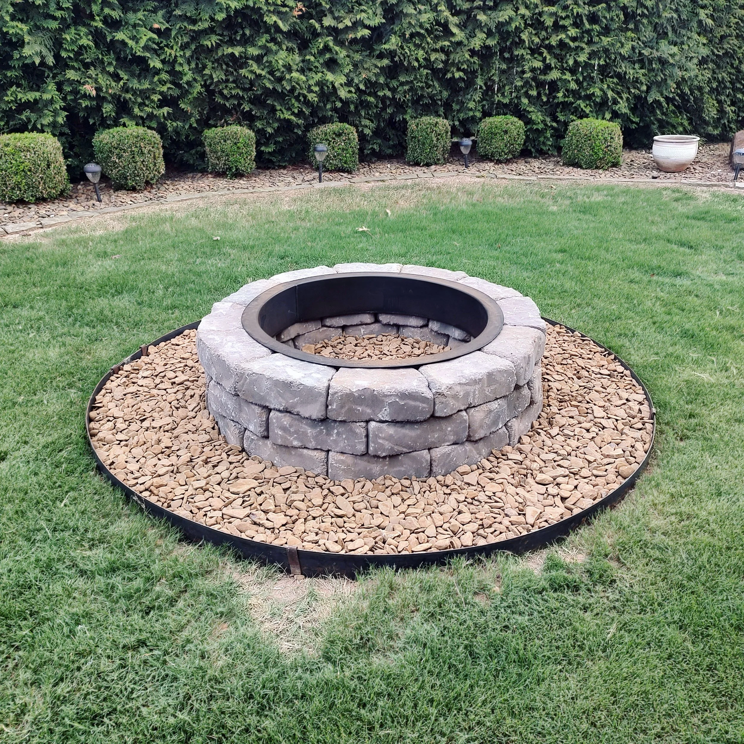 A circular fire pit made of gray stone bricks, surrounded by small tan rocks, set in a well-maintained grassy yard with trimmed bushes and trees in the background.