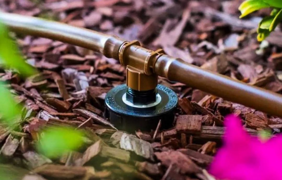 A garden sprinkler head with a brown plastic tube attached, installed in a mulched garden bed.